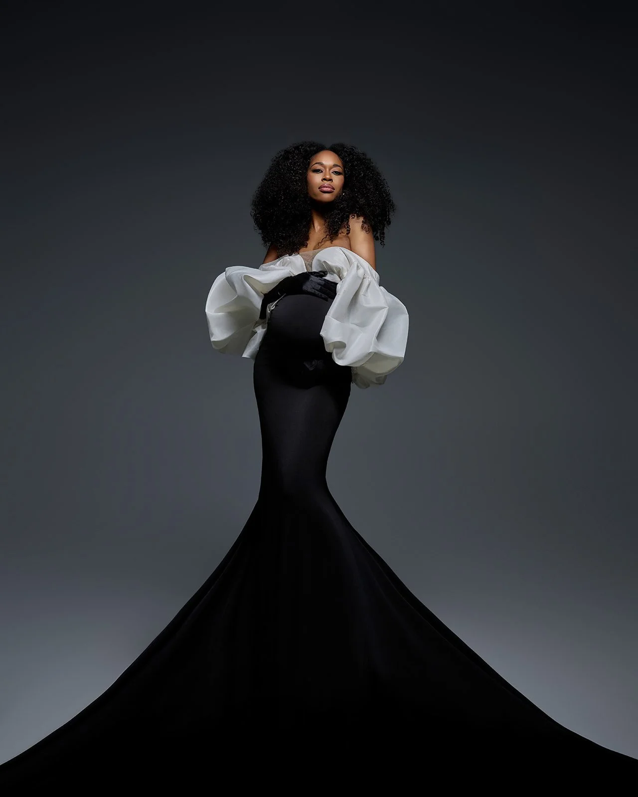 Sculptural maternity portrait of a pregnant woman wearing a black mermaid gown with oversized white sleeves.