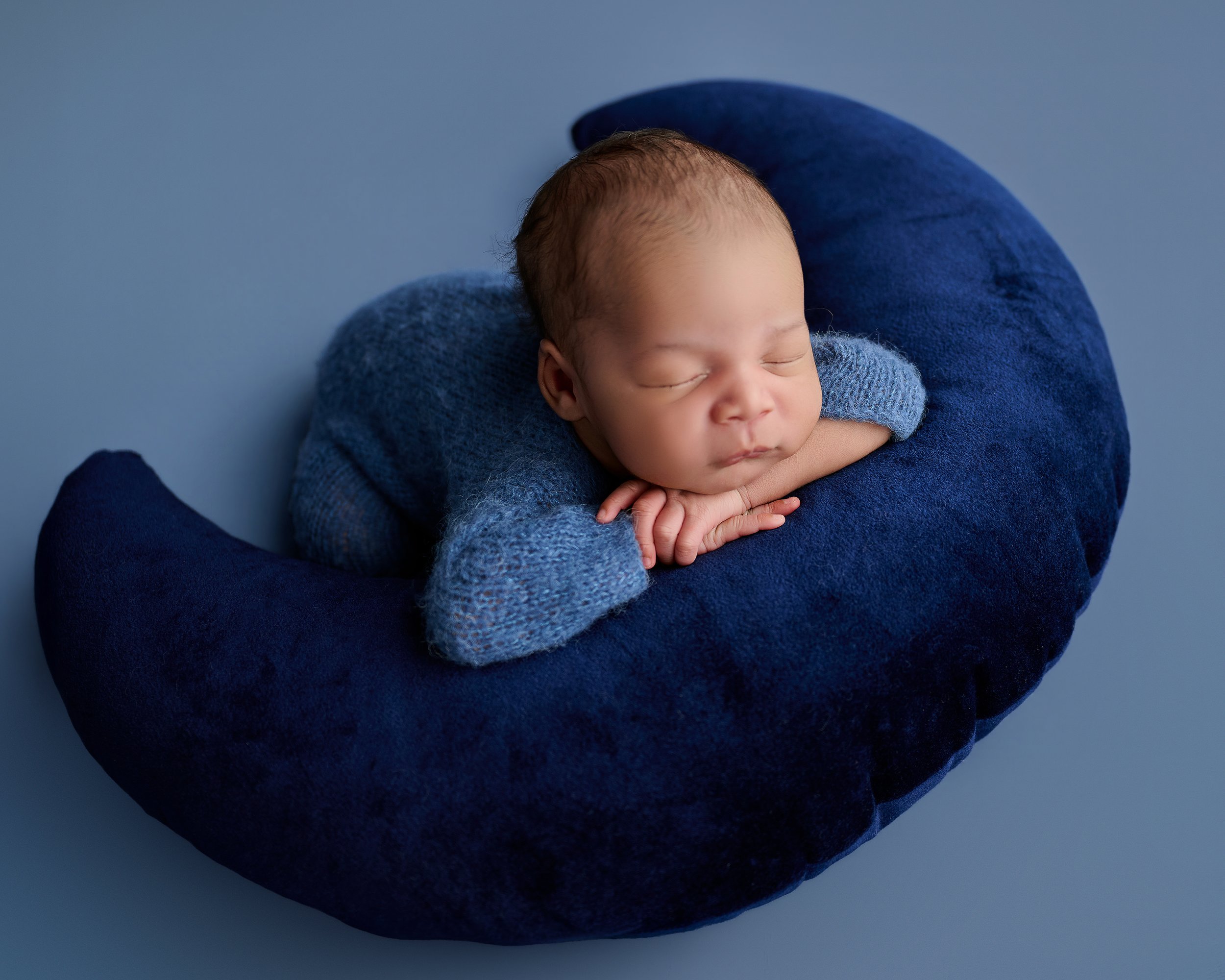 Fine art newborn portrait of a sleeping baby resting on a navy crescent moon prop in a soft blue color palette.