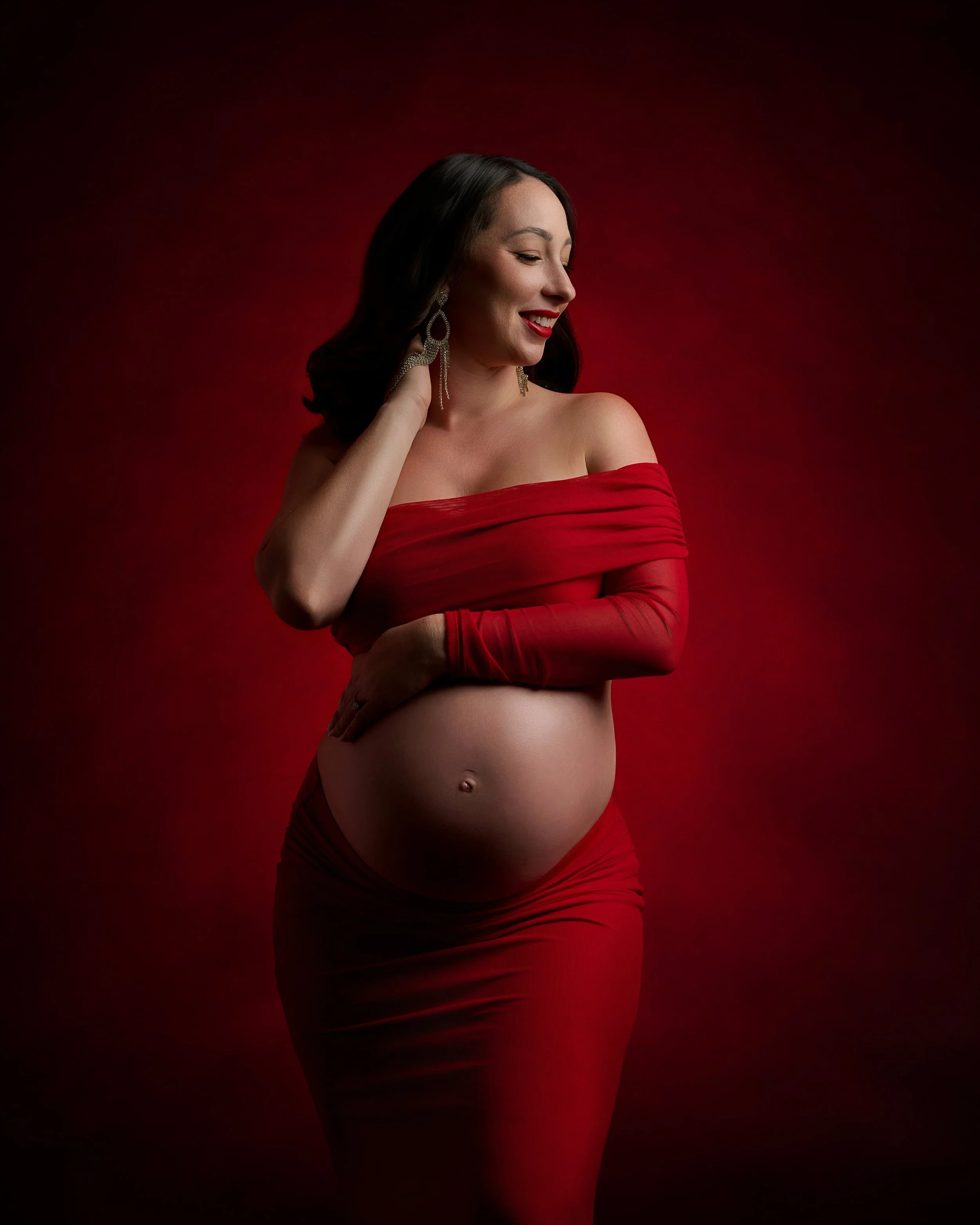 Pregnant woman wearing a red off-the-shoulder outfit against a deep red background in a dramatic Houston maternity portrait.