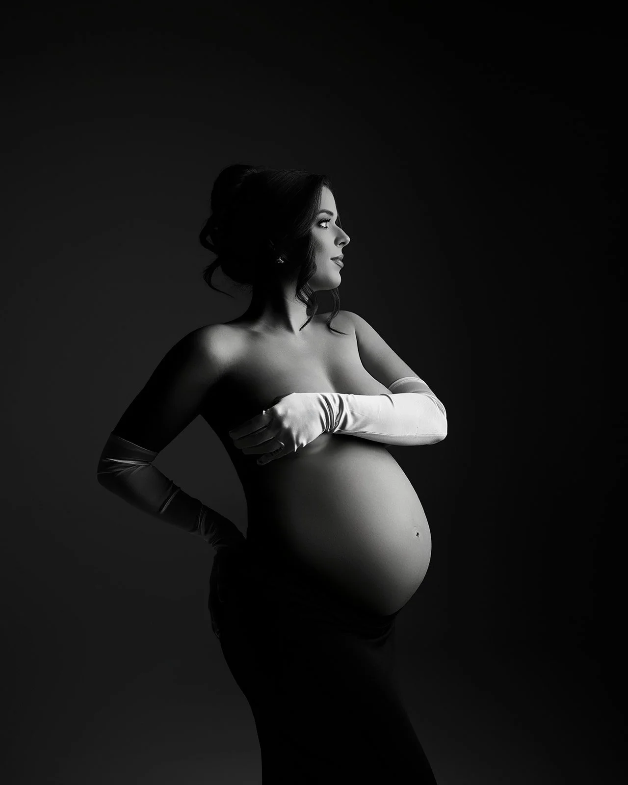 Black and white studio maternity portrait of a pregnant woman wearing long white gloves with dramatic sculpted lighting.