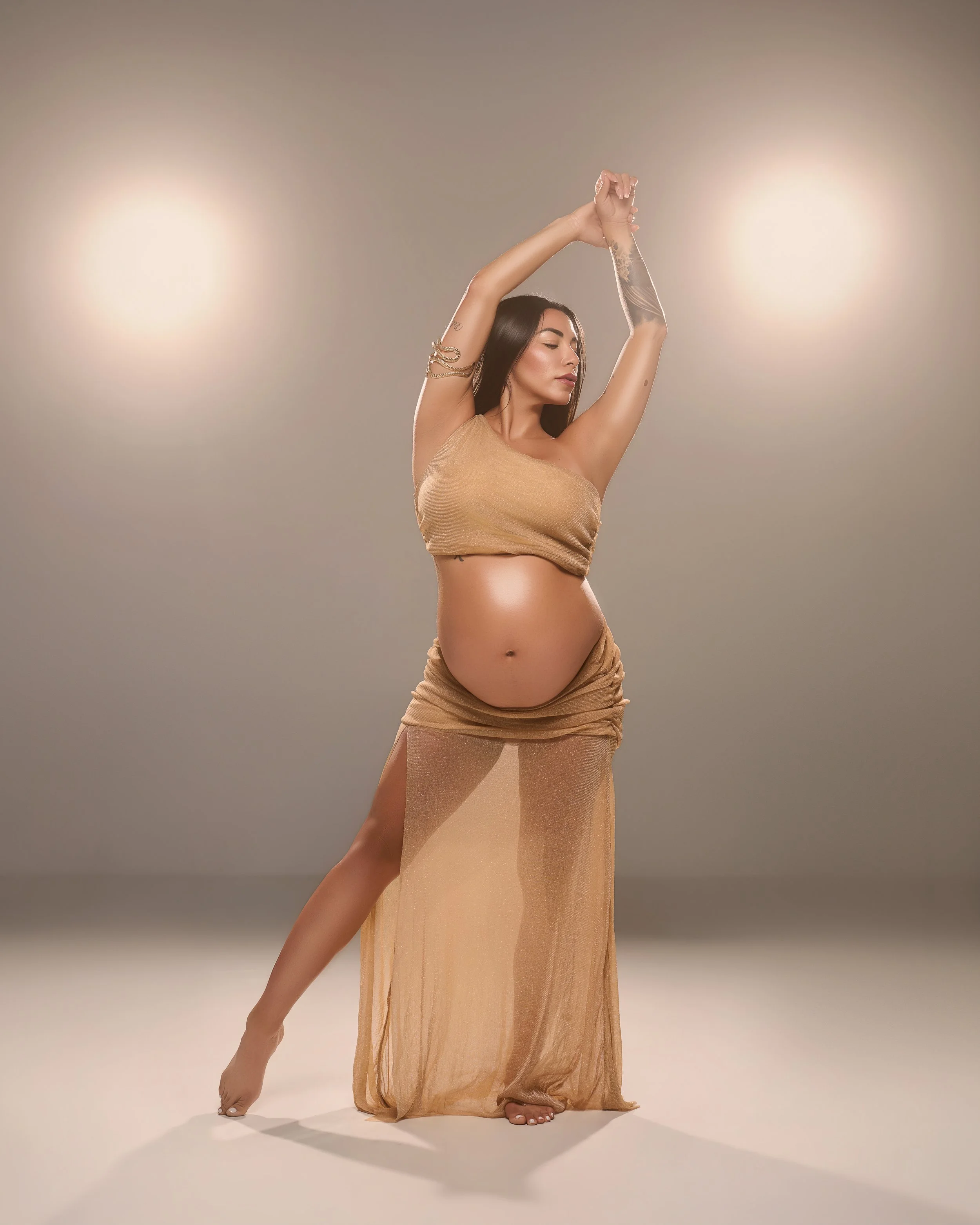 Pregnant woman in a sheer gold two-piece look posing between two warm lights during an editorial maternity session in Houston.