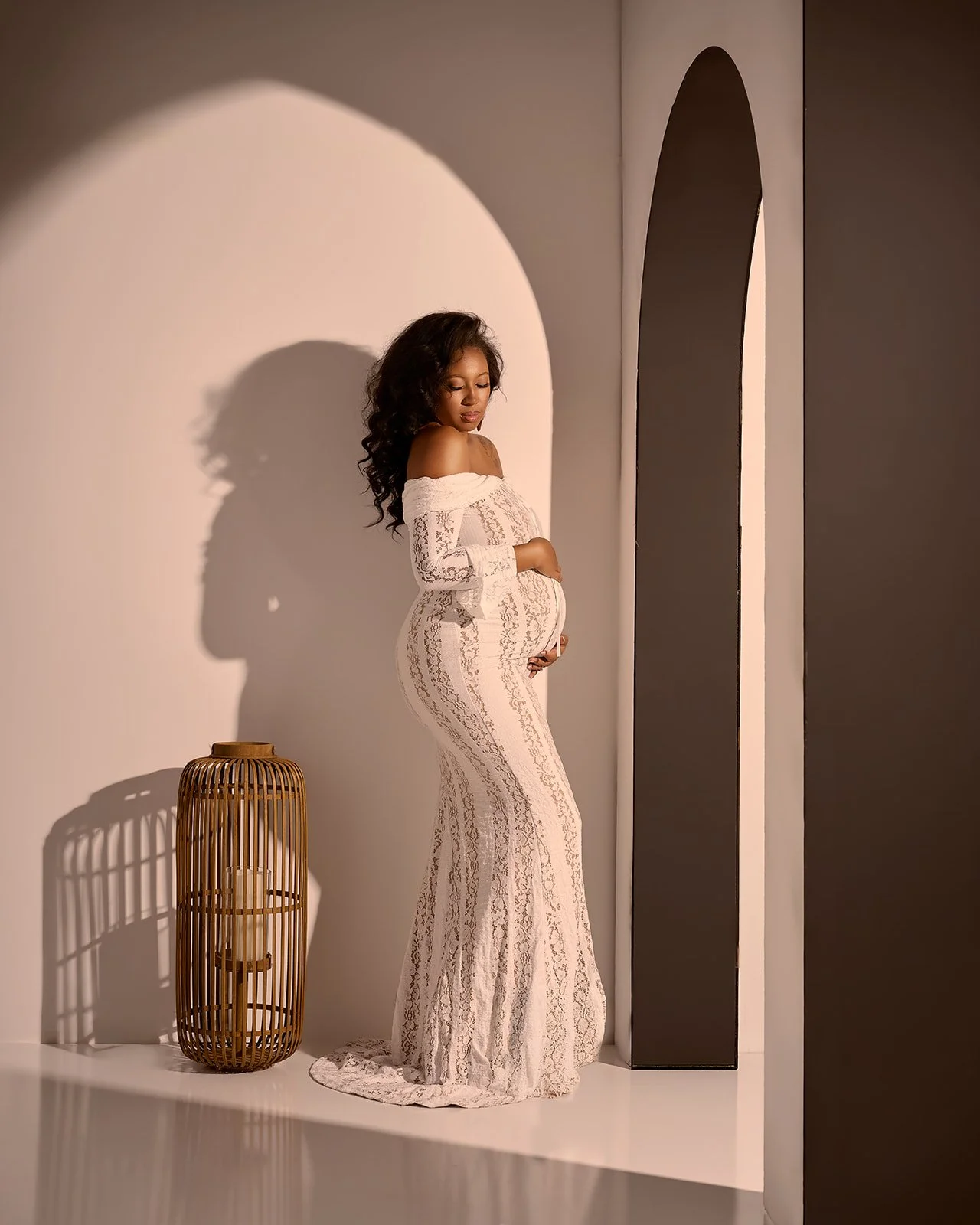 Pregnant woman wearing a fitted white lace gown posed beside an architectural arch with warm dramatic studio lighting.