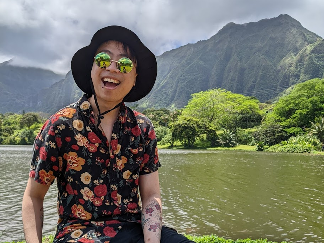 A smiling person wearing sunglasses, a black hat, and a floral shirt, sitting by a river with lush green trees and mountains in the background.