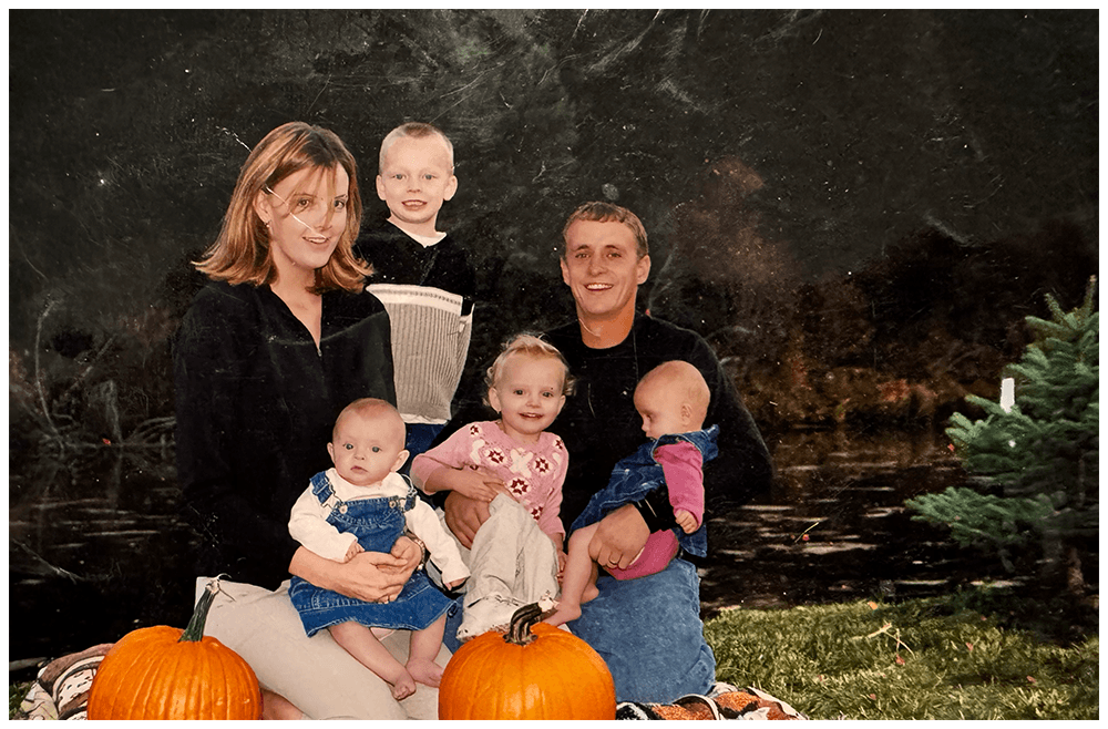 A family of six sitting outdoors at night with pumpkins and a small Christmas tree, smiling for the camera.