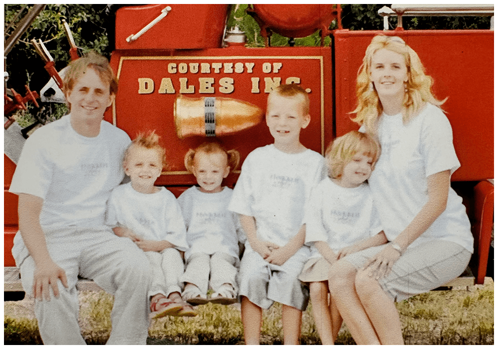 Family of six sitting on a firetruck with the sign 'Courtesy of Dales Inc.' in the background.