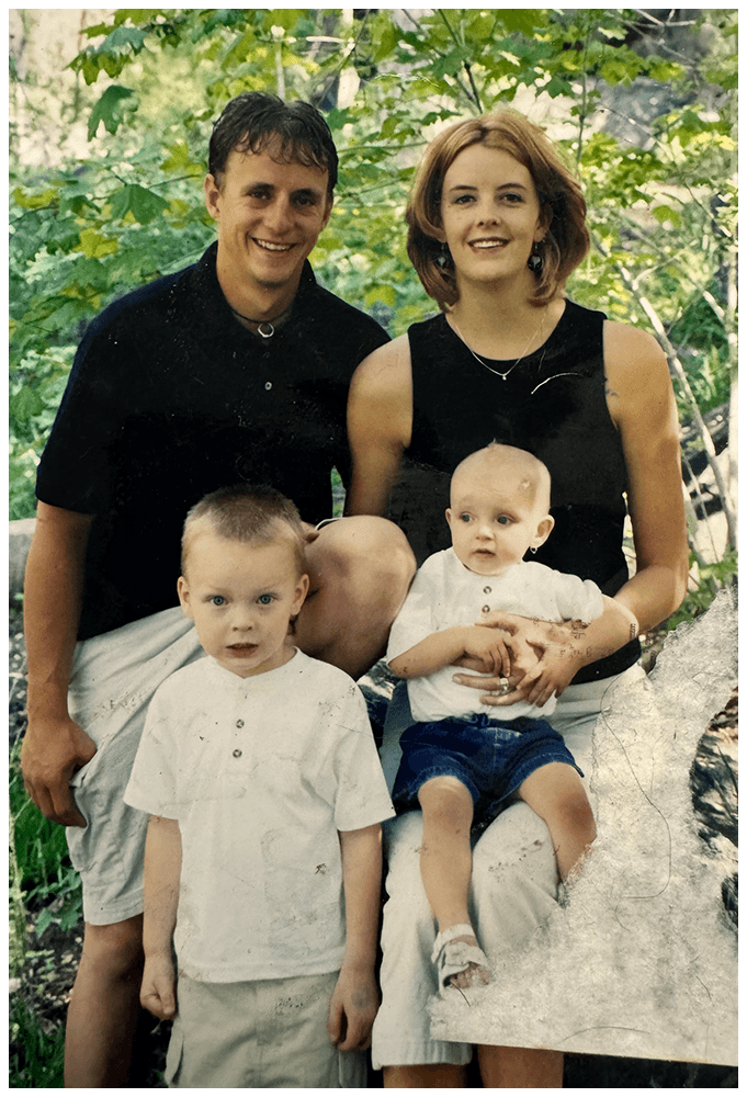 A family photo of two adults and two young boys outdoors among green trees. The adults are smiling, and the boys are sitting on a large rock, with one looking at the camera and the other looking slightly away.