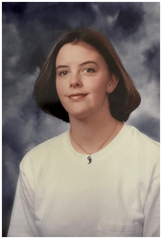 A woman with shoulder-length brown hair, wearing a white shirt and a necklace with a yin-yang pendant, poses in front of a cloudy, dark background.