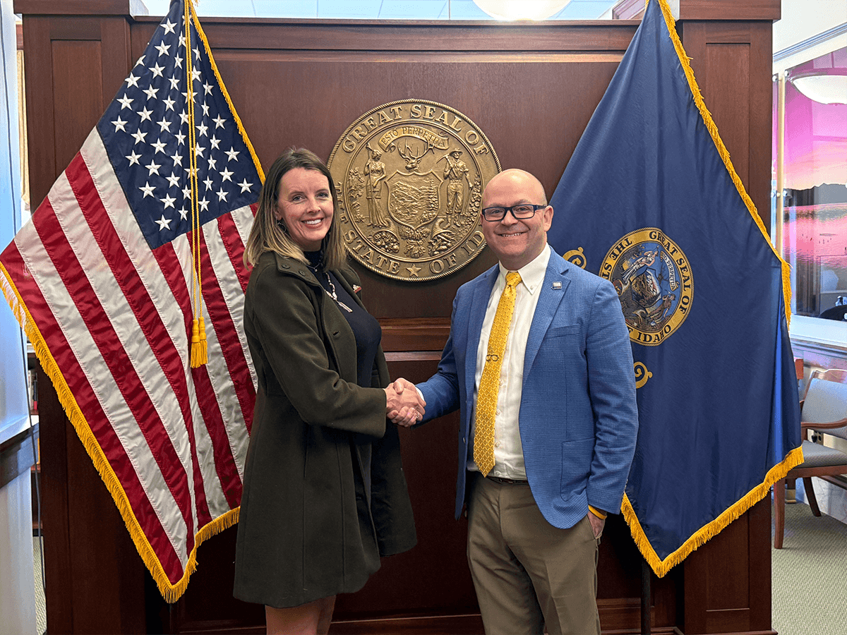 A woman and a man shake hands in front of a wooden wall with the Great Seal of the State of Idaho behind them. The woman is wearing a dark coat and the man is wearing a blue suit with a yellow tie. There are American and Idaho state flags on either side of them.