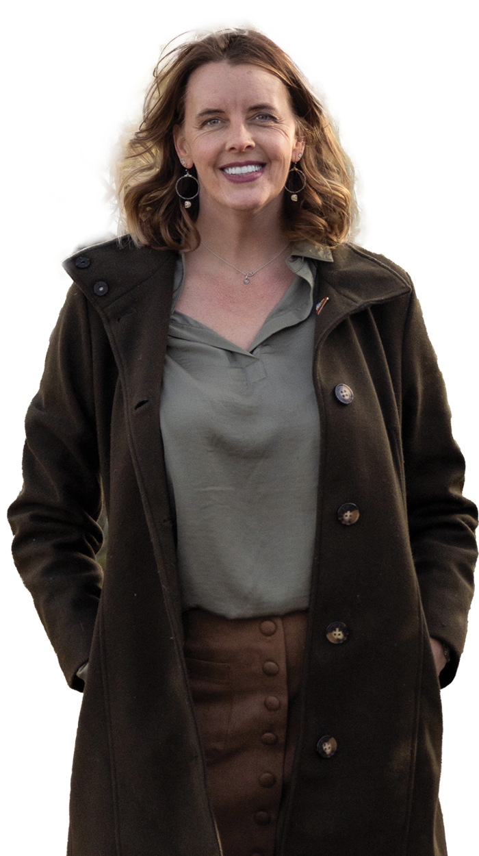 A woman with shoulder-length wavy hair smiling, wearing a dark coat over a gray blouse and brown skirt, standing outdoors.