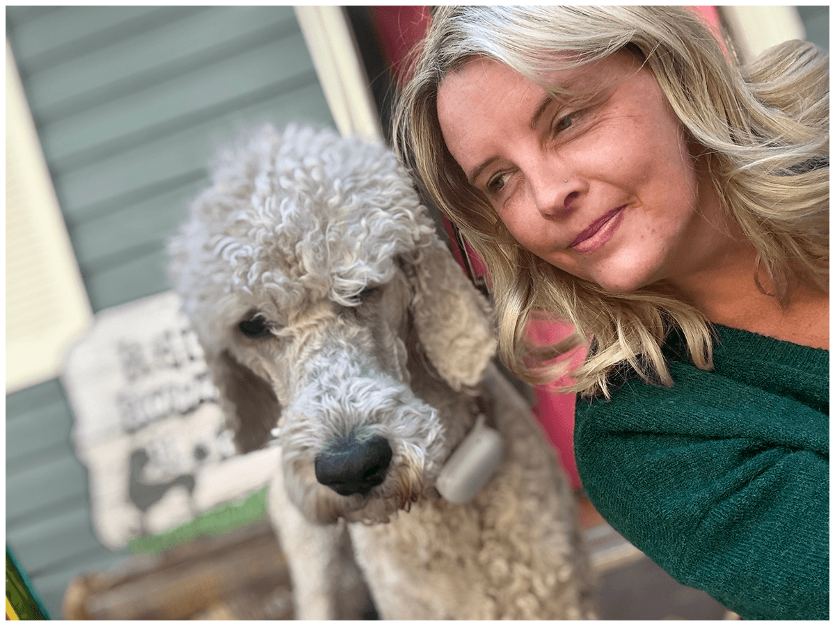 A woman with blonde hair taking a selfie with a curly-haired dog, likely a poodle. They are outside near a house with green siding and a decorative dog-themed sign in the background.