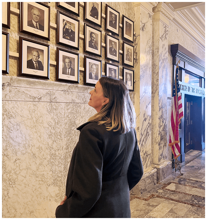 A woman with shoulder-length hair looking at framed black-and-white portraits of older men on a marble wall inside a government building with an American flag to the right.