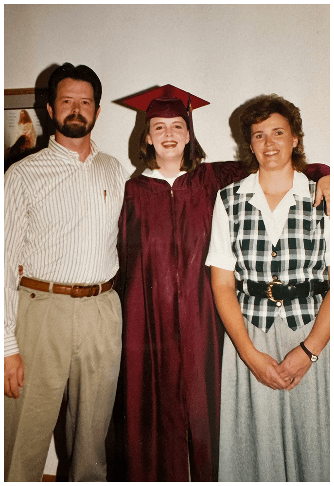 A young woman in a maroon graduation gown and cap stands between an older man in a striped shirt and a woman in a checkered blouse. All three are smiling, with the woman in graduation attire having her arms around the other two. The background is a plain wall with a framed picture.