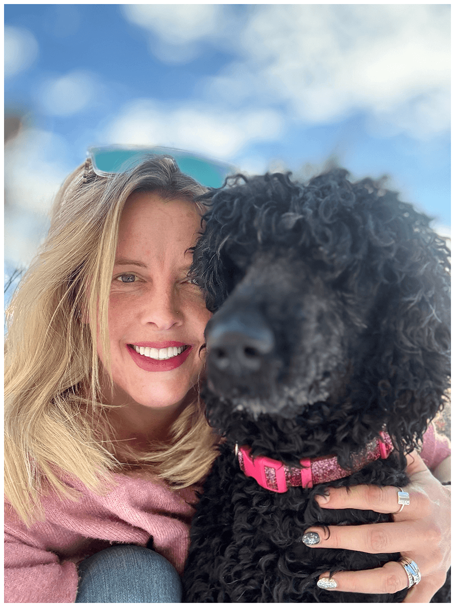 Smiling woman with blonde hair and sunglasses on her head holding a curly black dog with a pink collar, outdoor sky background.