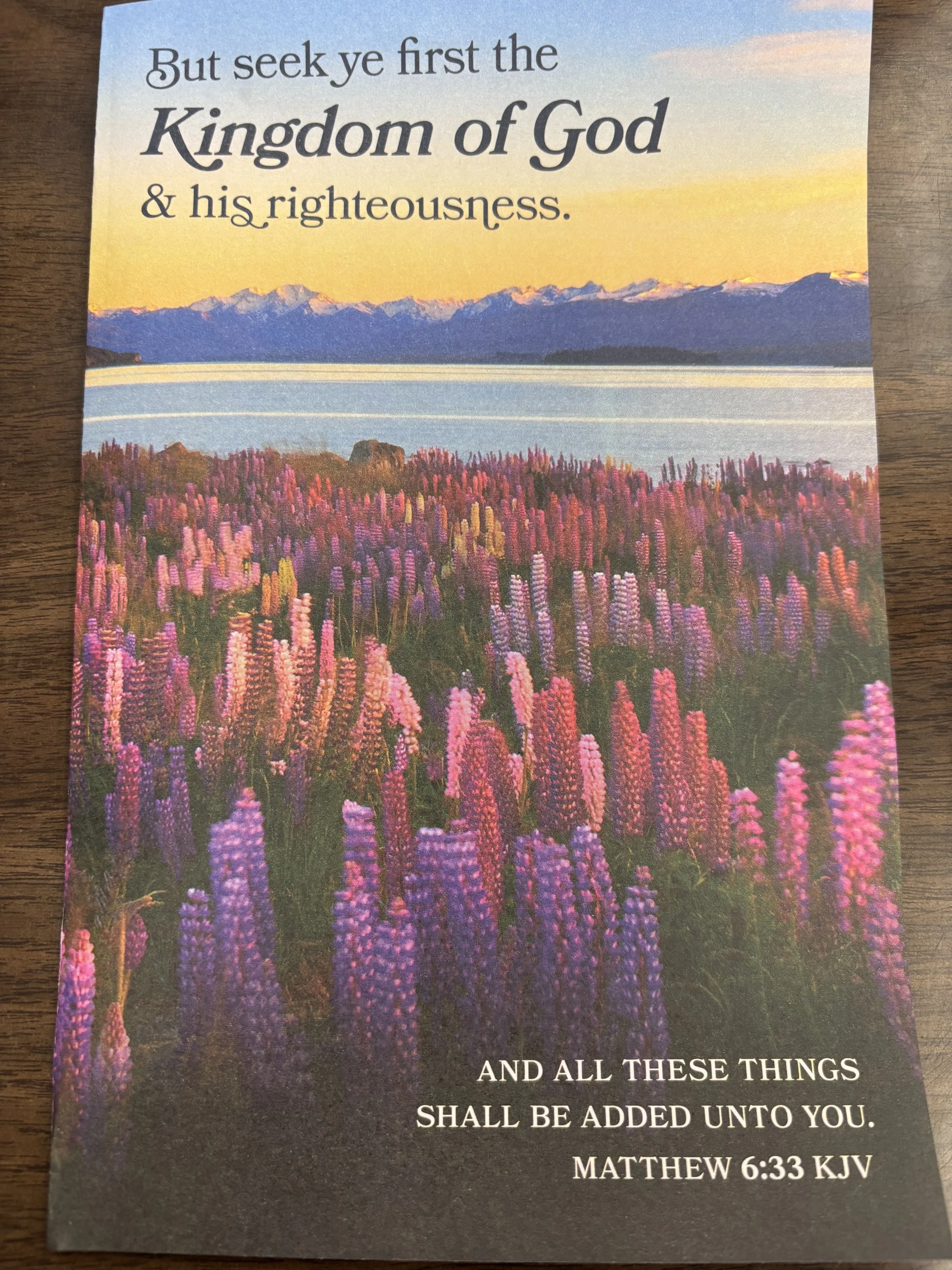 A scenic landscape of purple flowers in the foreground, a lake, mountains in the background, and a yellow and blue sky at sunset or sunrise.