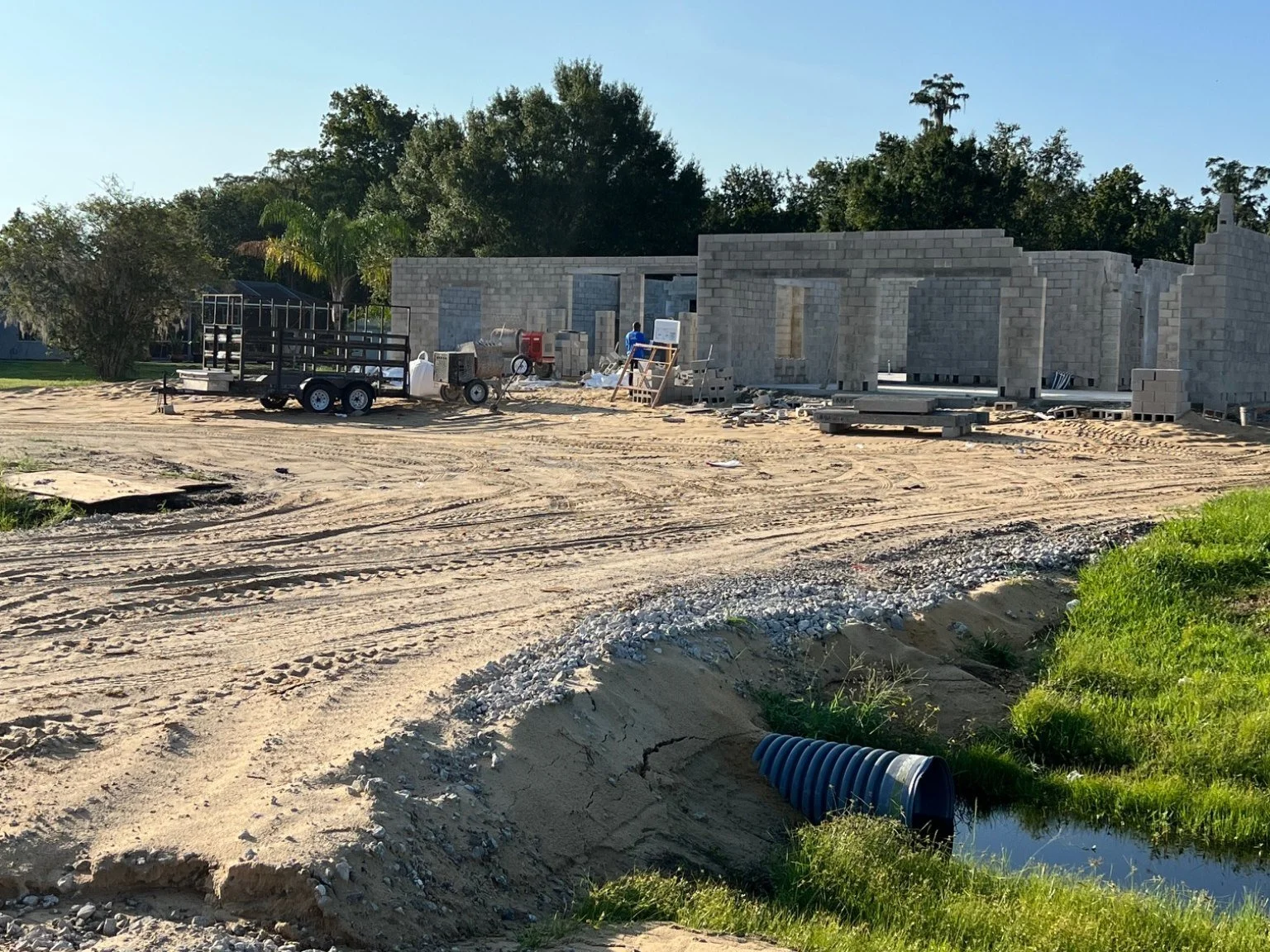 Construction site with partially built concrete block house, construction workers, a trailer, equipment, and dirt ground, with trees in the background and a blue sky.