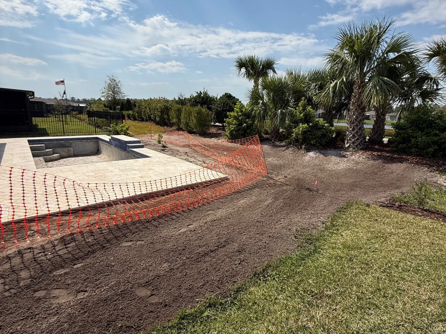 Backyard in construction with a swimming pool, palm trees, bushes, and orange safety fencing, on a sunny day with partly cloudy skies.