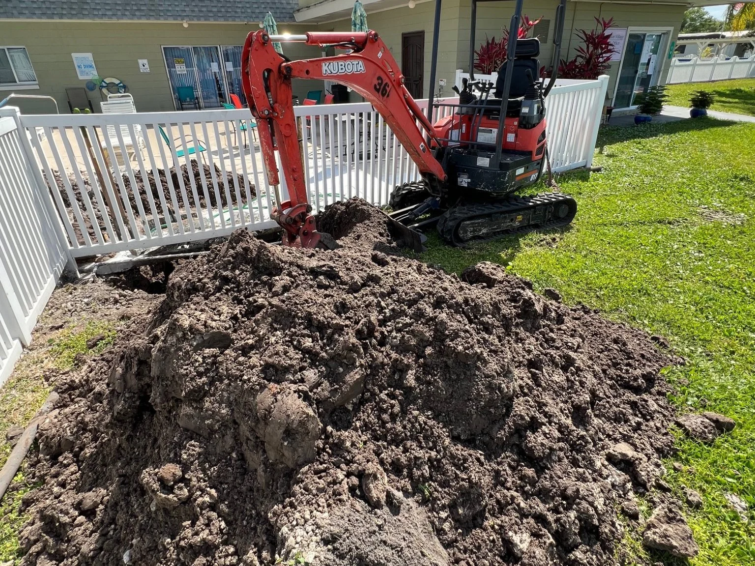 A red Kubota mini excavator digging a hole near a white fence in a backyard. There is a large pile of dark dirt and rocks in the foreground. The background shows a patio area with chairs and tables, and a beige house with windows and sliding doors.