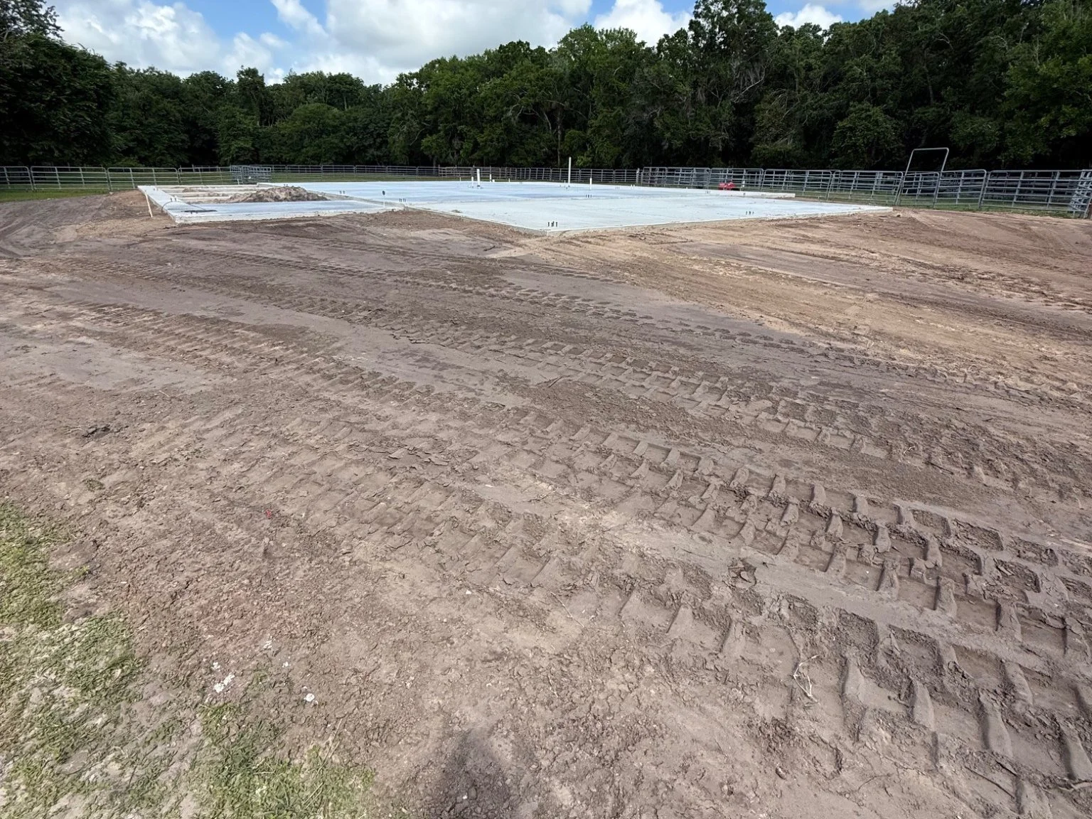 Construction site with cleared dirt and tracks leading to a concrete foundation with fencing, surrounded by trees.