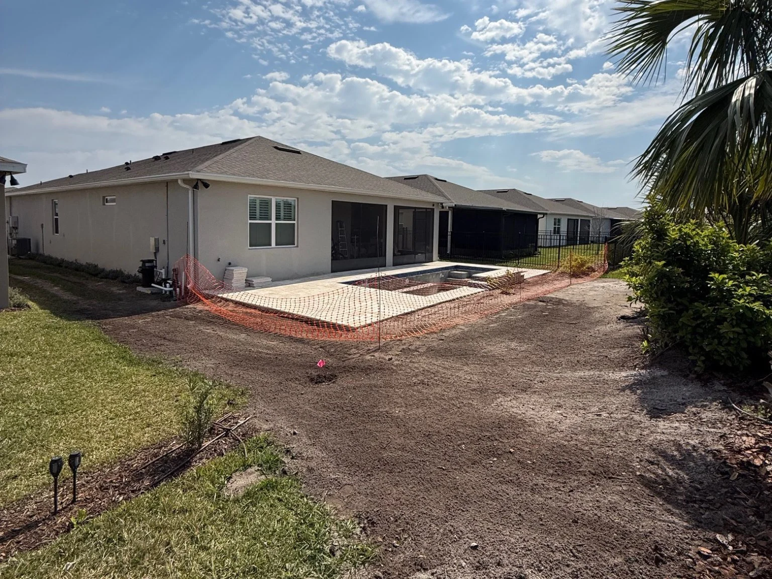 Backyard under construction with a poured concrete pad, surrounded by orange safety fencing, adjacent to a beige house with screened porch, against a partly cloudy sky.