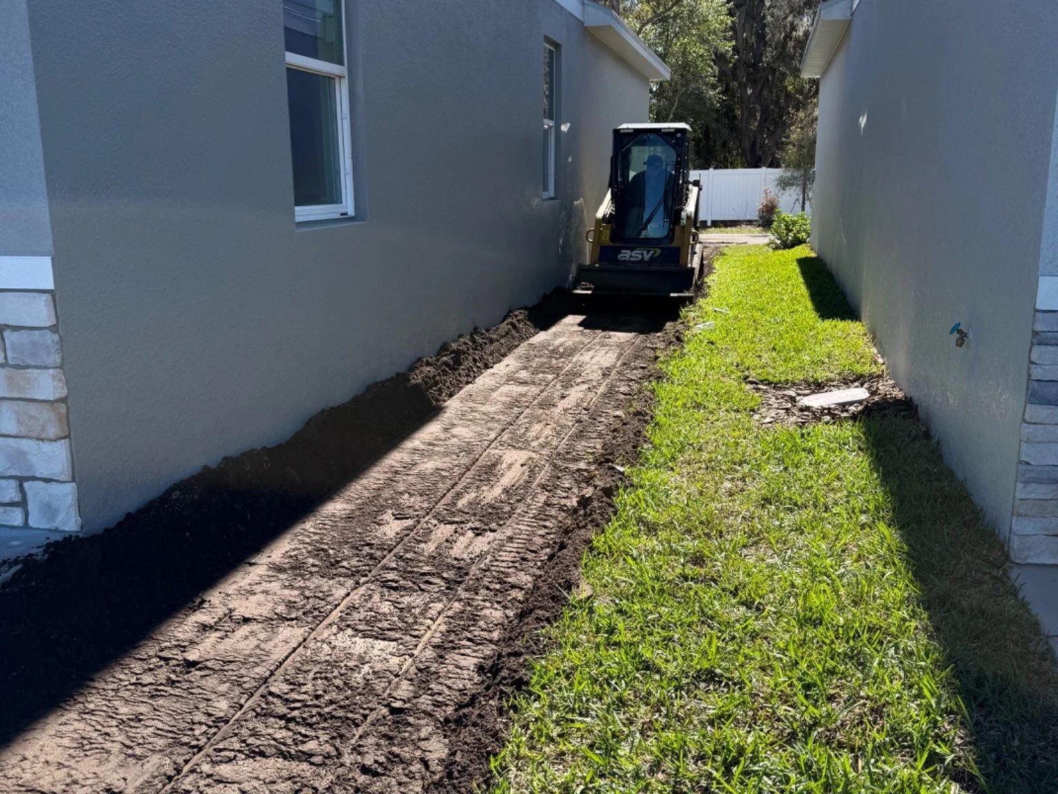 A small skid-steer loader working on a narrow strip of land between two houses, preparing the ground for paving or landscaping.