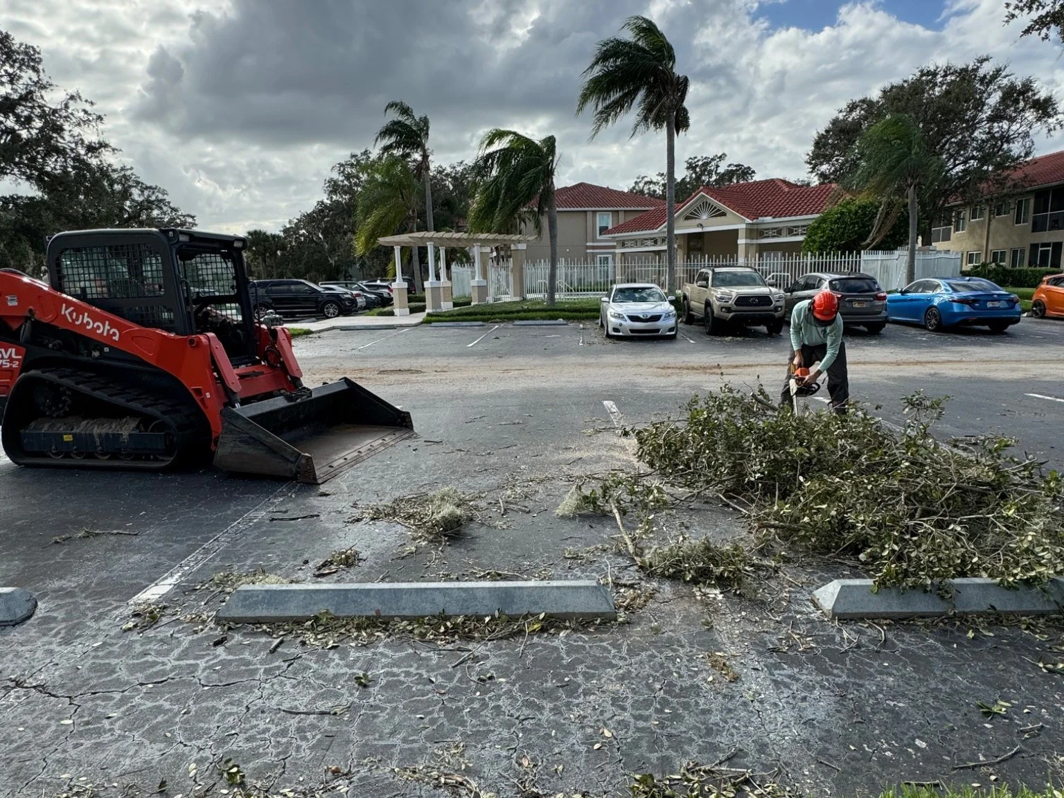 A parking lot where a worker wearing a hard hat is trimming bushes with a power tool. A small red excavator is nearby, and there are several parked cars in the background.