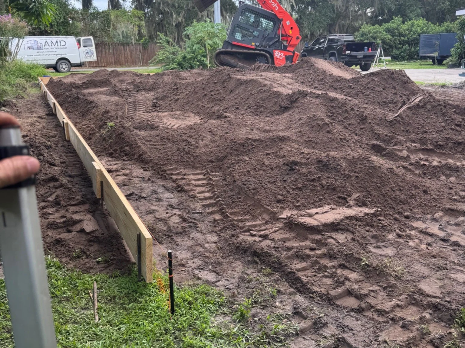 A construction site with a wooden form frame in place for pouring concrete, surrounded by piles of dirt and tracked tire marks, with a small red skid-steer loader in the background.