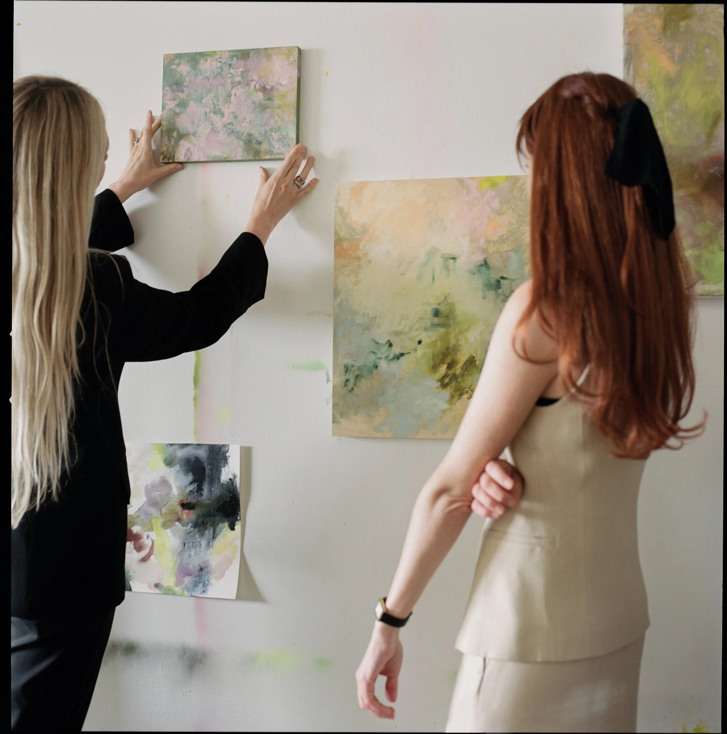 Two women looking at and arranging abstract paintings on a white wall in an art gallery or studio.