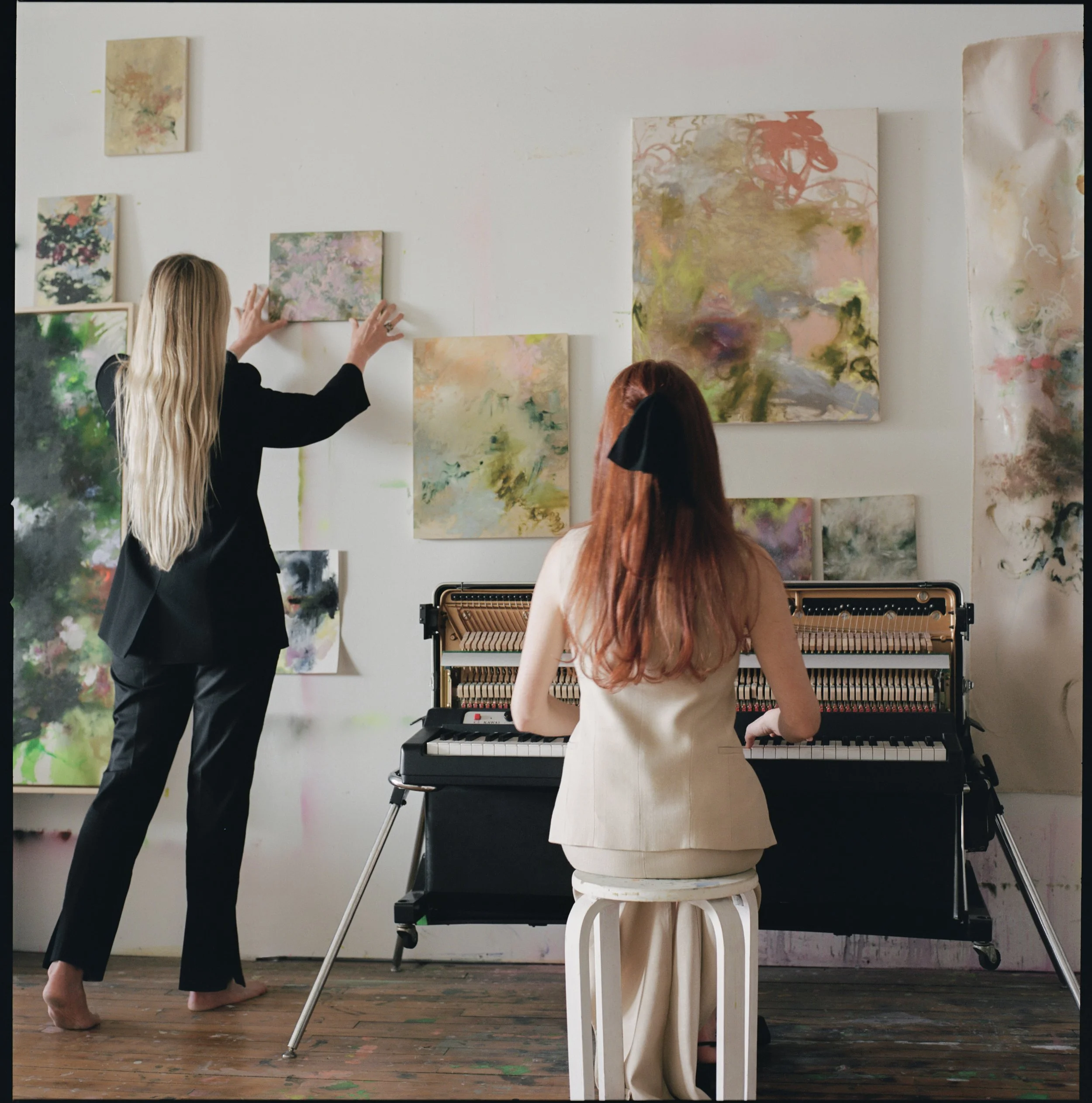 Two women in an art studio. One standing and adjusting abstract paintings on the wall, the other sitting on a stool playing an upright piano.