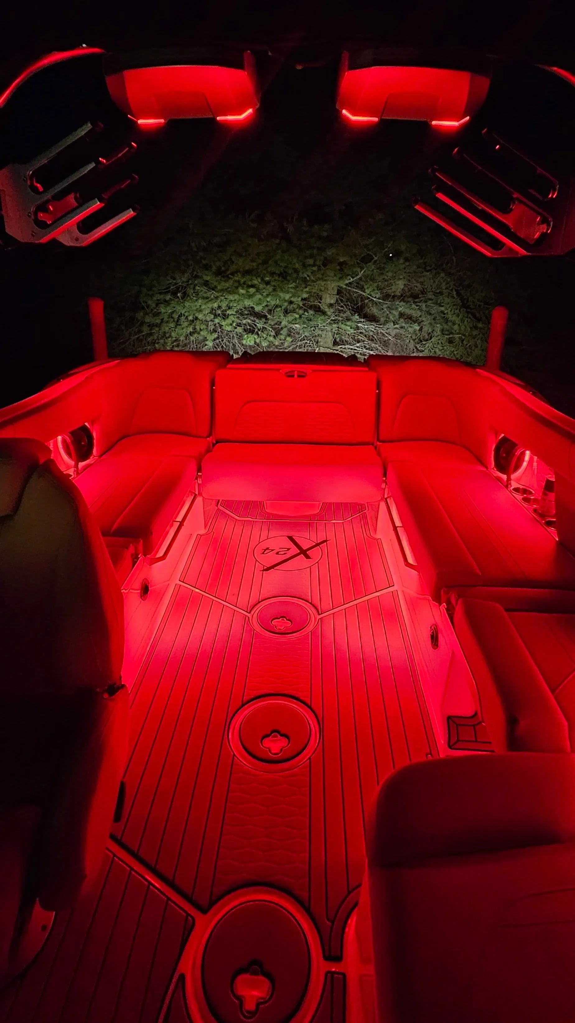 The interior of a vehicle illuminated with red lights, featuring comfortable red leather seating arranged in a U-shape and a textured floor mat with the Mercedes-Benz logo and the number 24.
