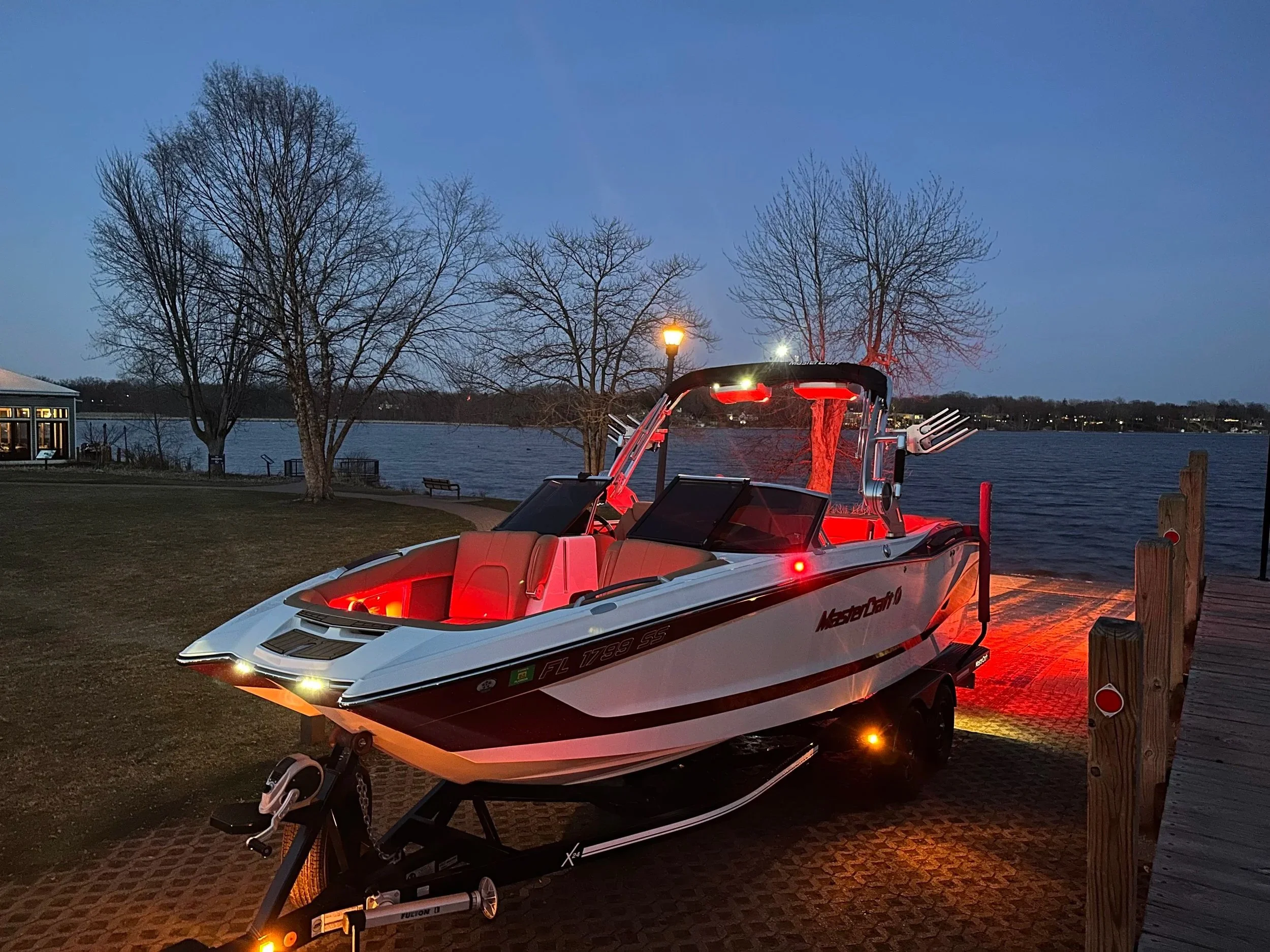 A white and red motorboat on a trailer parked on a dock near a lake during dusk, with leafless trees and a lamppost in the background.
