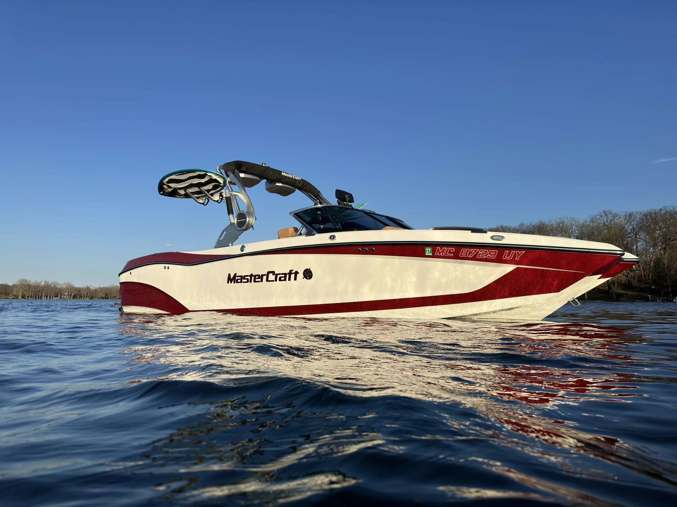 A white and red MasterCraft speedboat on calm water with a clear blue sky overhead and trees in the background.