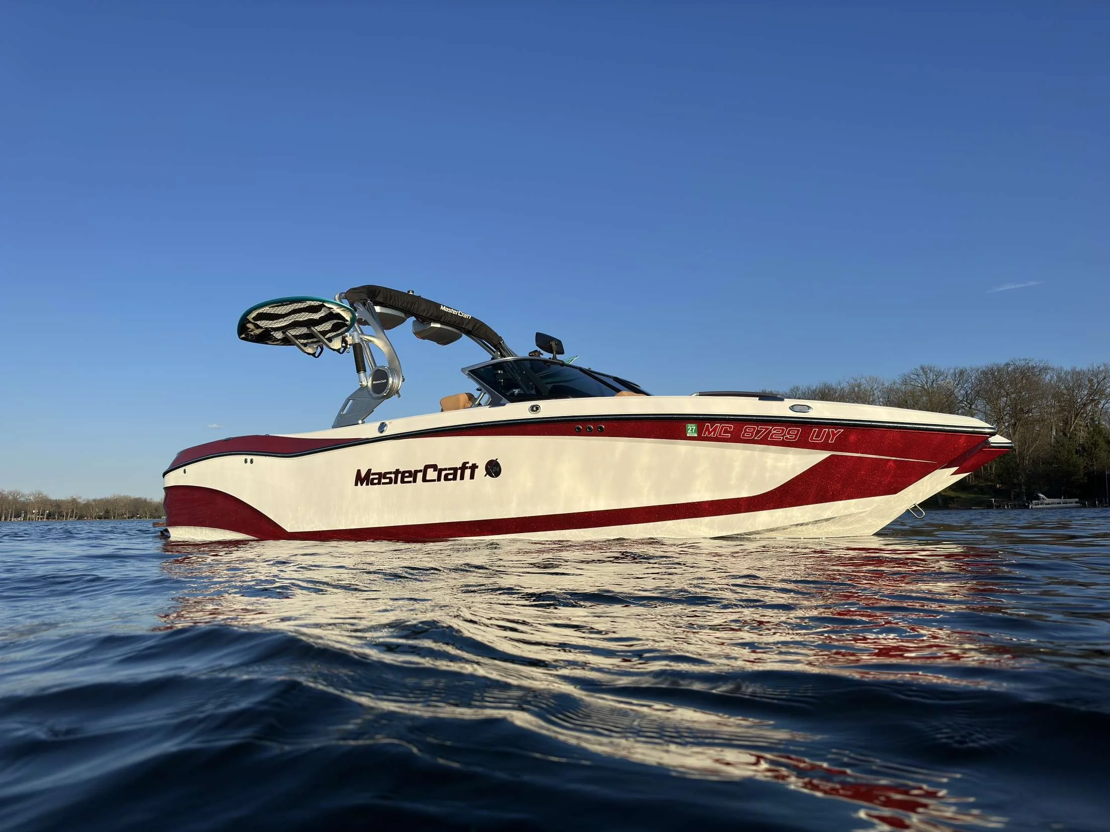 A white and red MasterCraft speedboat on calm water with a clear blue sky and trees in the background.