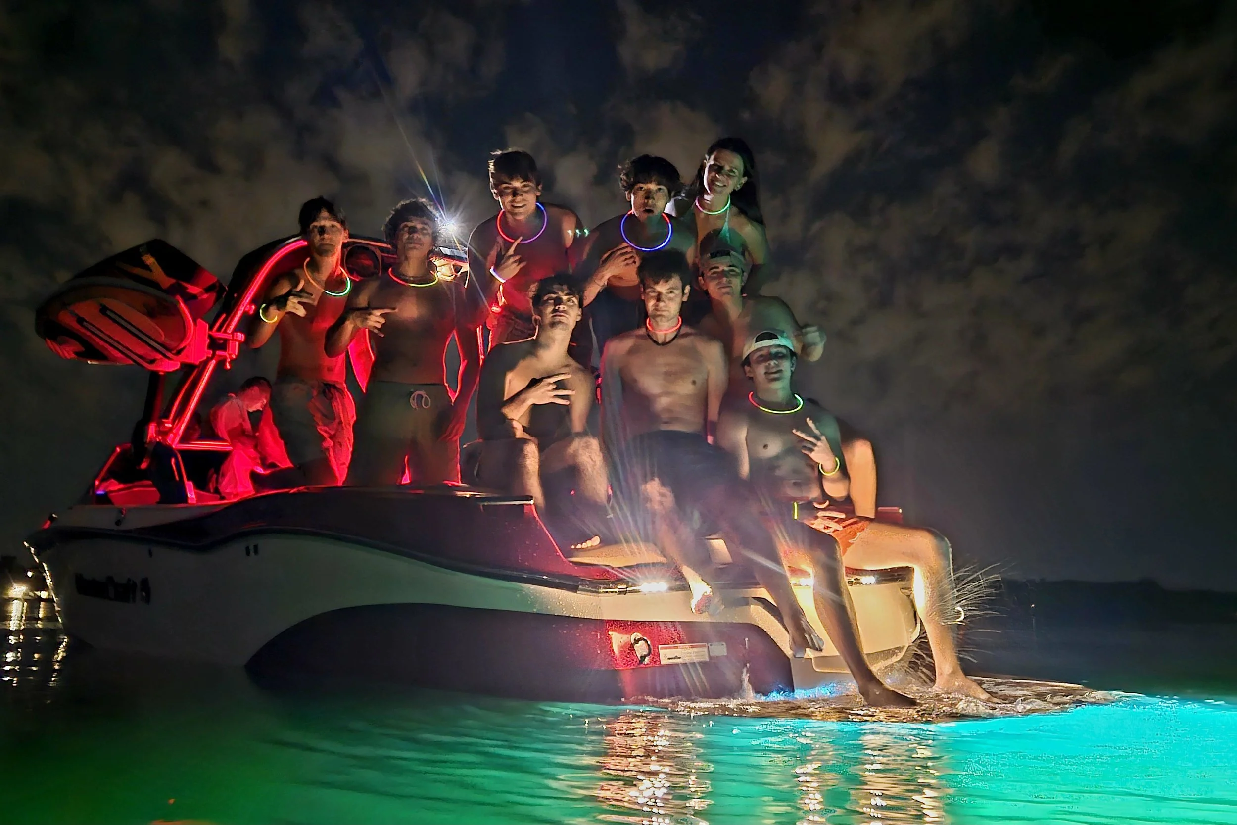 Nighttime photo of eleven young people on a boat, wearing glow sticks and necklaces, with some shirtless. They are smiling and making peace signs, with glowing water in the foreground and dark, cloudy sky in the background.