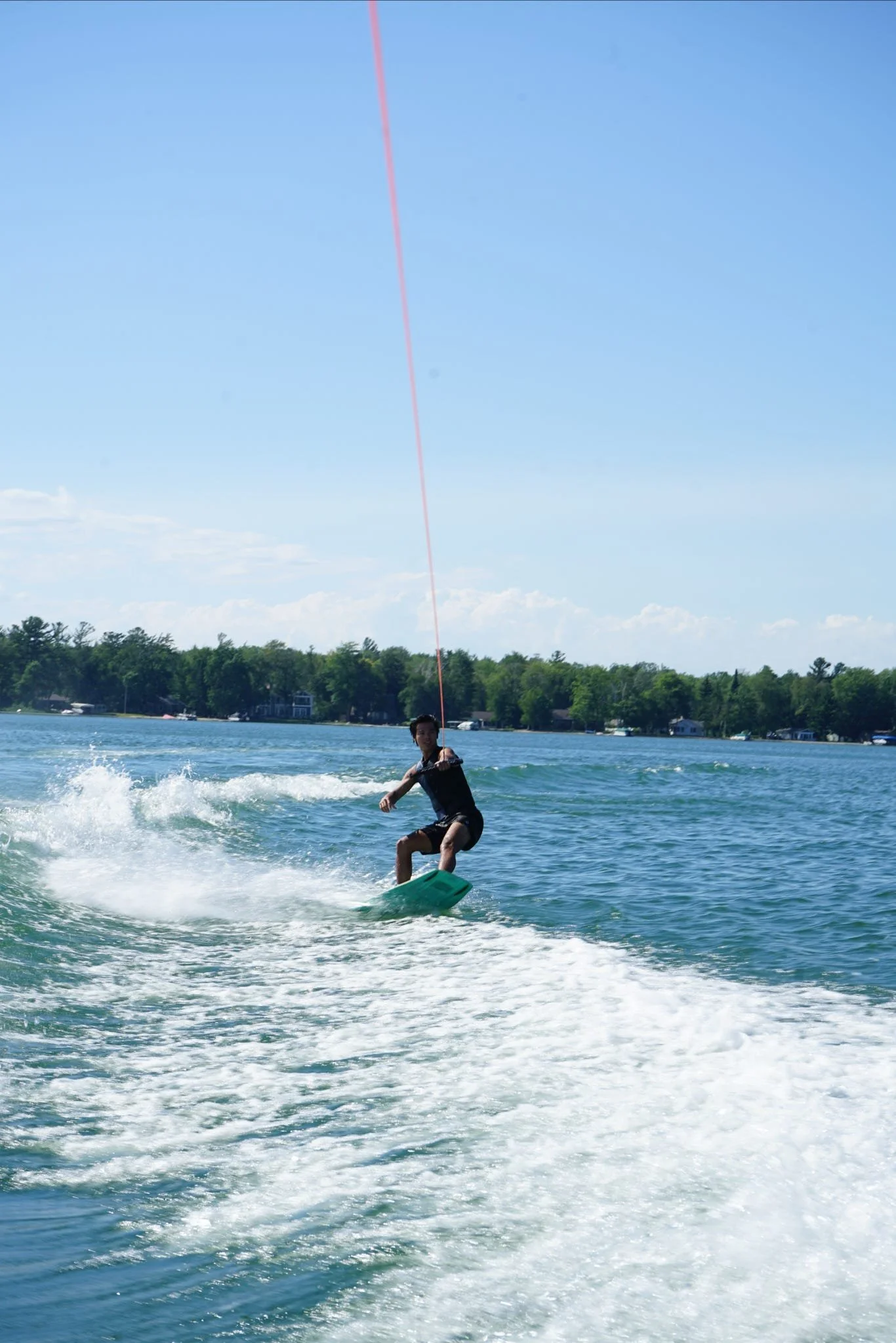 A person wakeboarding on a lake on a sunny day, with a line attached to a boat, and trees and houses in the background.