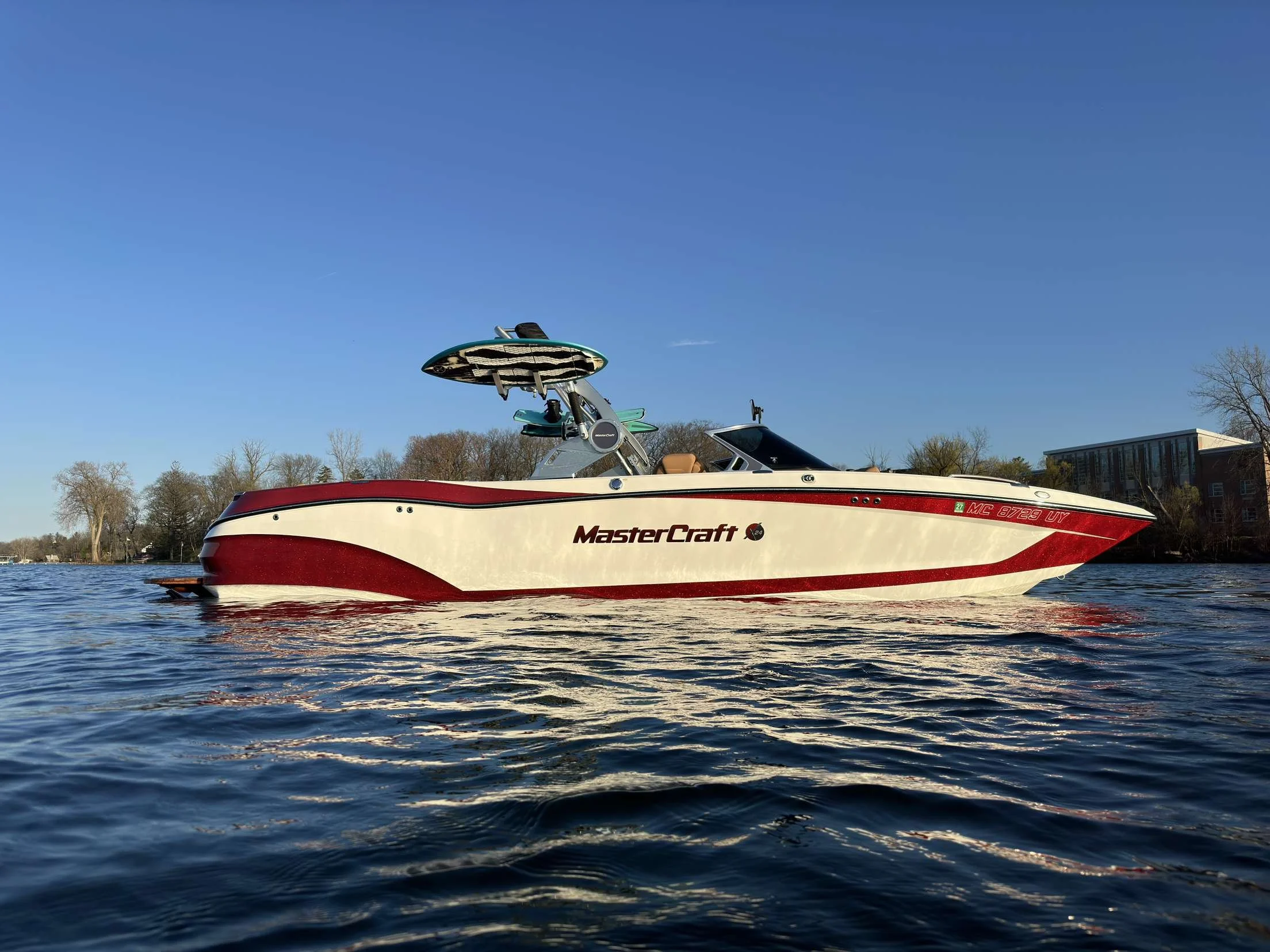 A white and red MasterCraft speedboat on a calm body of water with trees and a building in the background, under a clear blue sky.