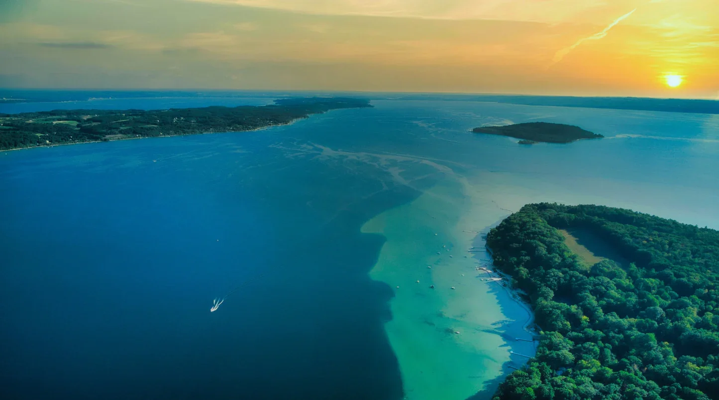Aerial view of a large lake at sunset with a small boat leaving a wake, surrounded by green forests and islands, under a partly cloudy sky.
