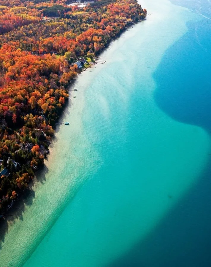Aerial view of a lake with clear blue water, a shoreline with trees in fall colors, and houses along the coast.