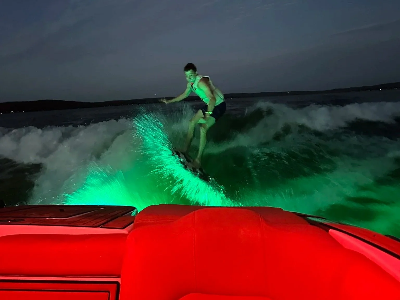 A person wakes on a wakeboard at night, riding on the water illuminated by green lights, as seen from the back of a boat with red seats.