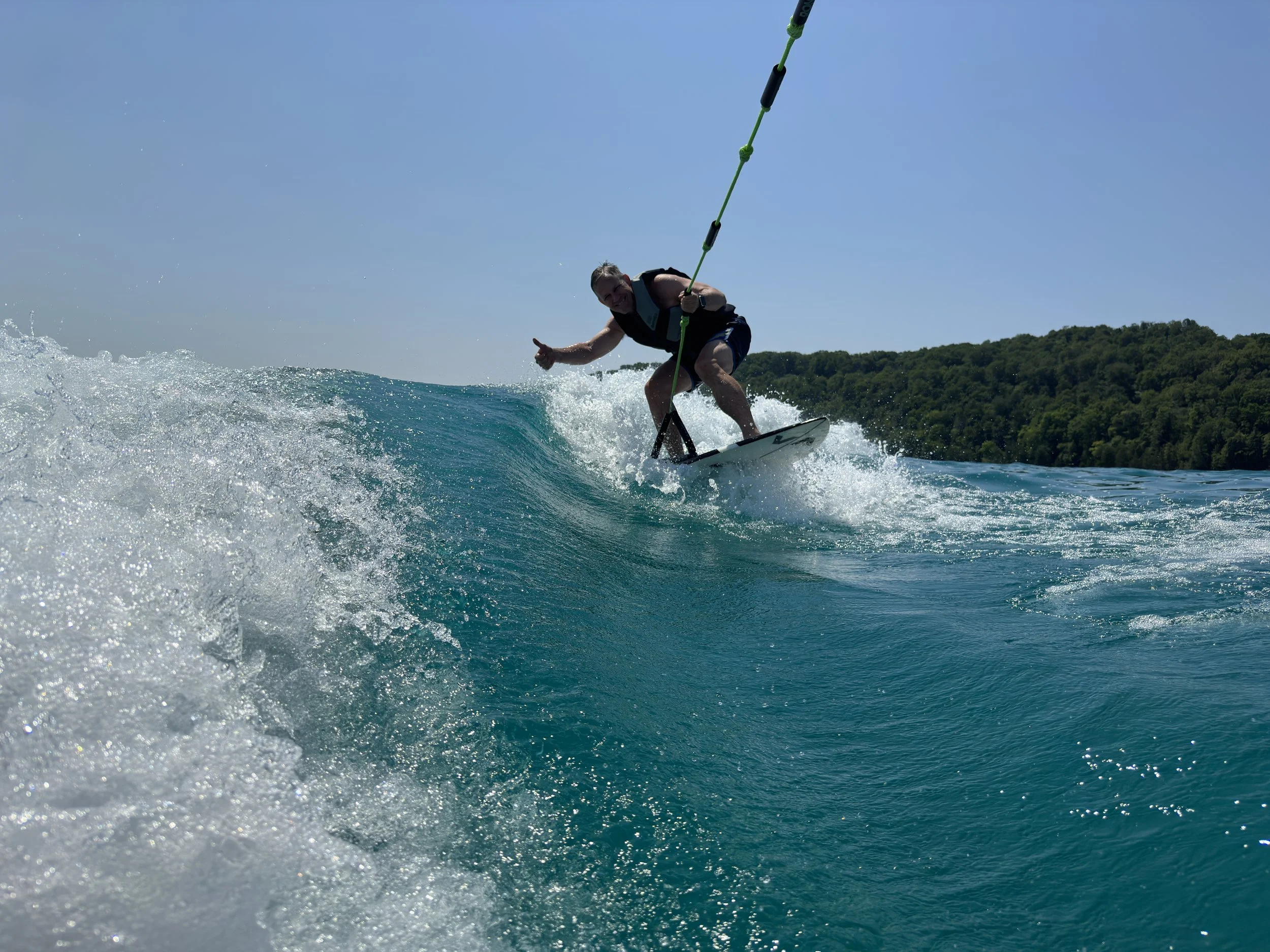 Person wakeboarding on the water with a green rope, leaning back and giving a thumbs up, head slightly down, with a distant tree-covered shoreline in the background.