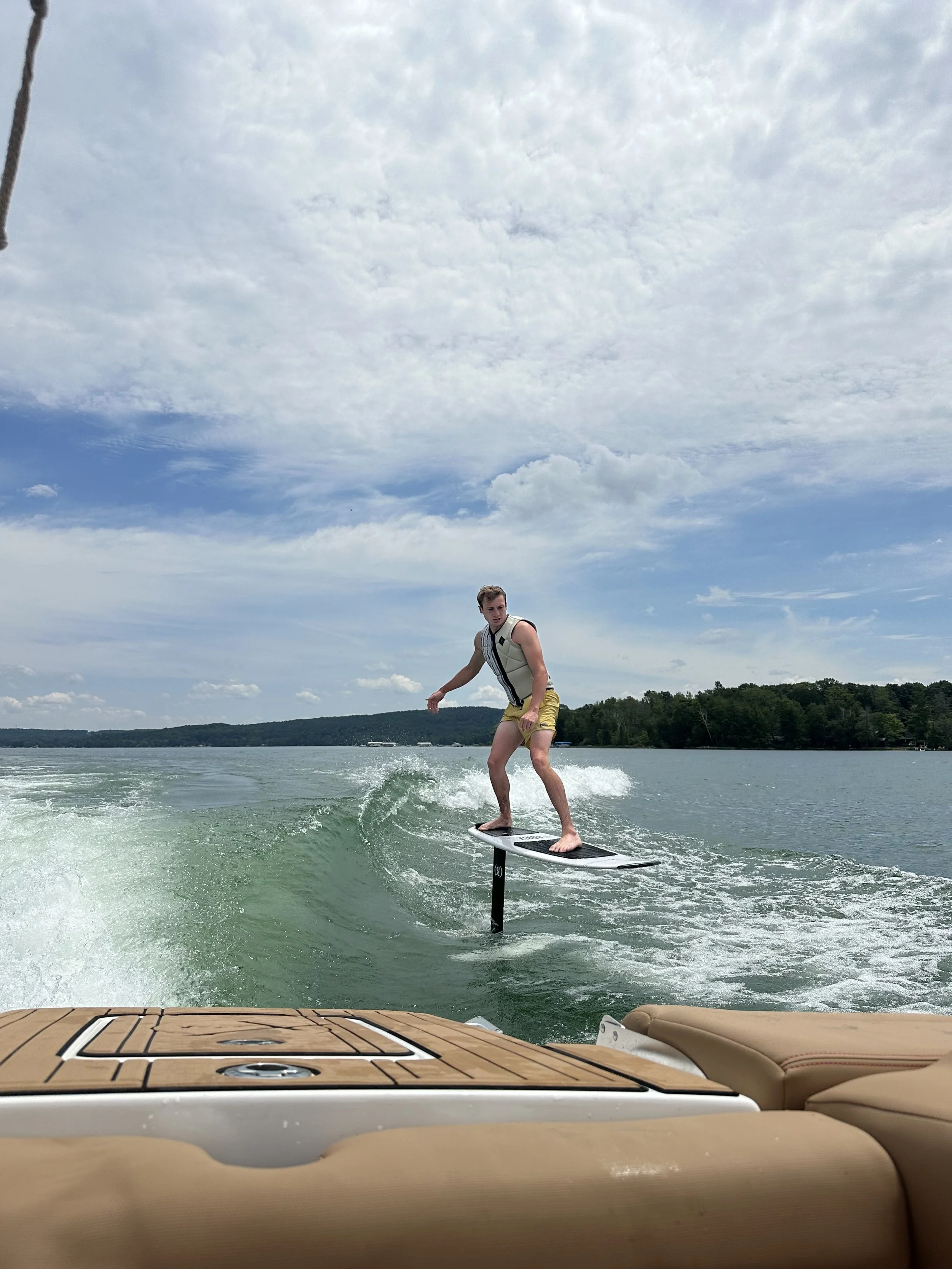 Person riding a hydrofoil wakeboard on a lake during a partly cloudy day, with trees in the background.