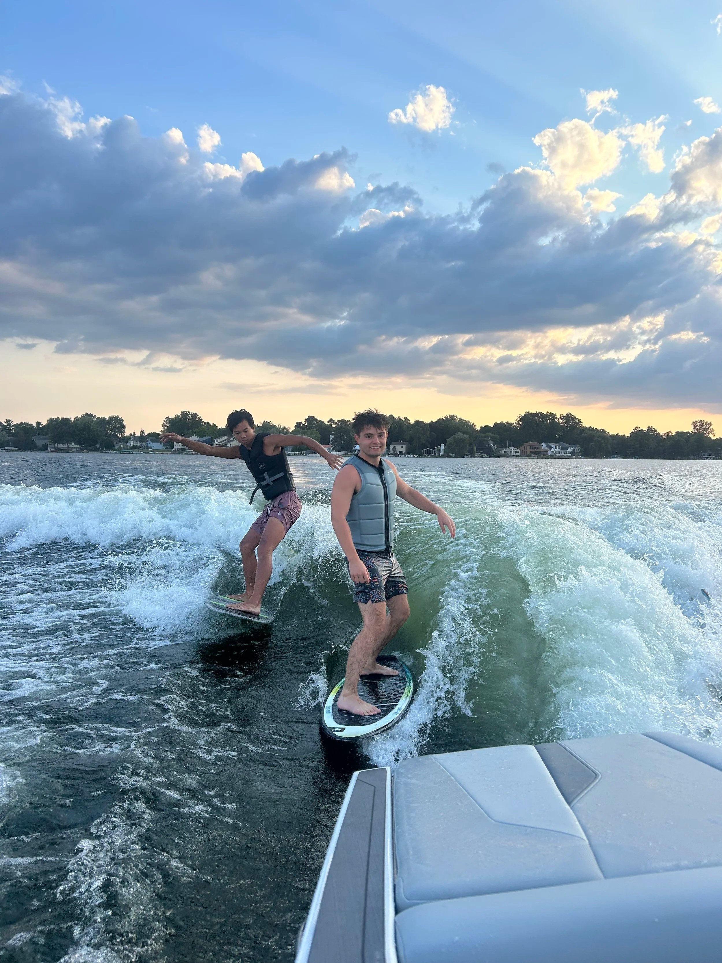 Two young men wakeboarding on a lake during sunset, with a house-lined shoreline in the background and a mostly cloudy sky overhead.