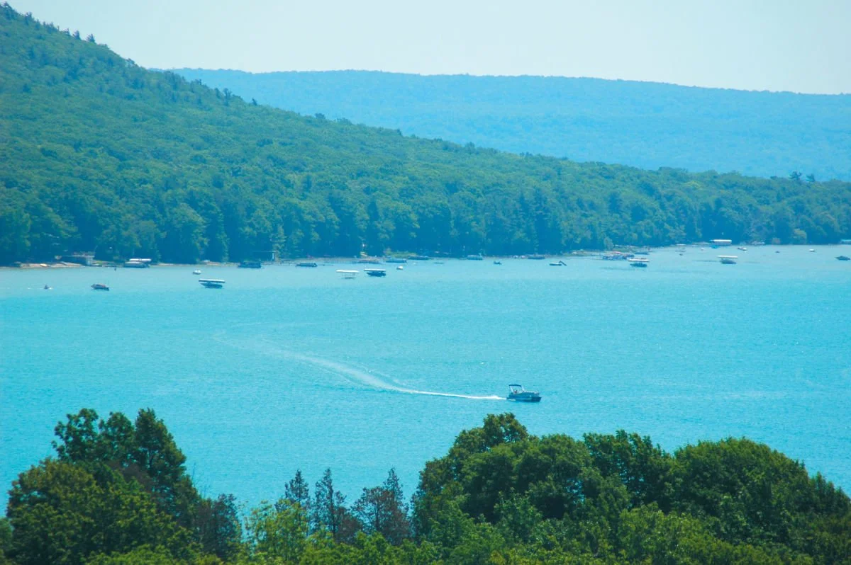 A large body of water with boats and a motorboat leaving a wake, surrounded by lush green hills and distant mountains.