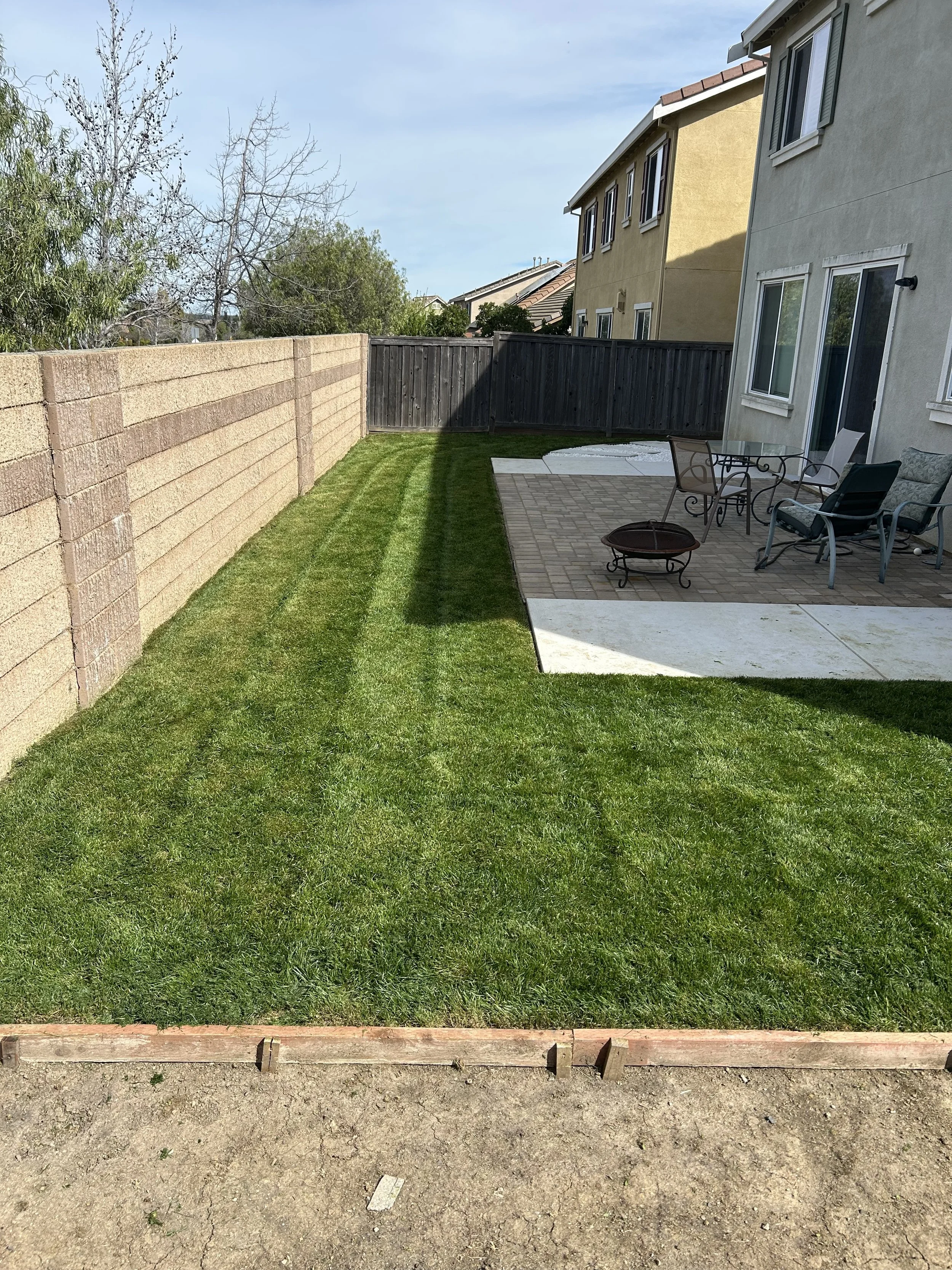 Backyard with freshly laid green grass, a brick patio with outdoor furniture, and a wooden fence.