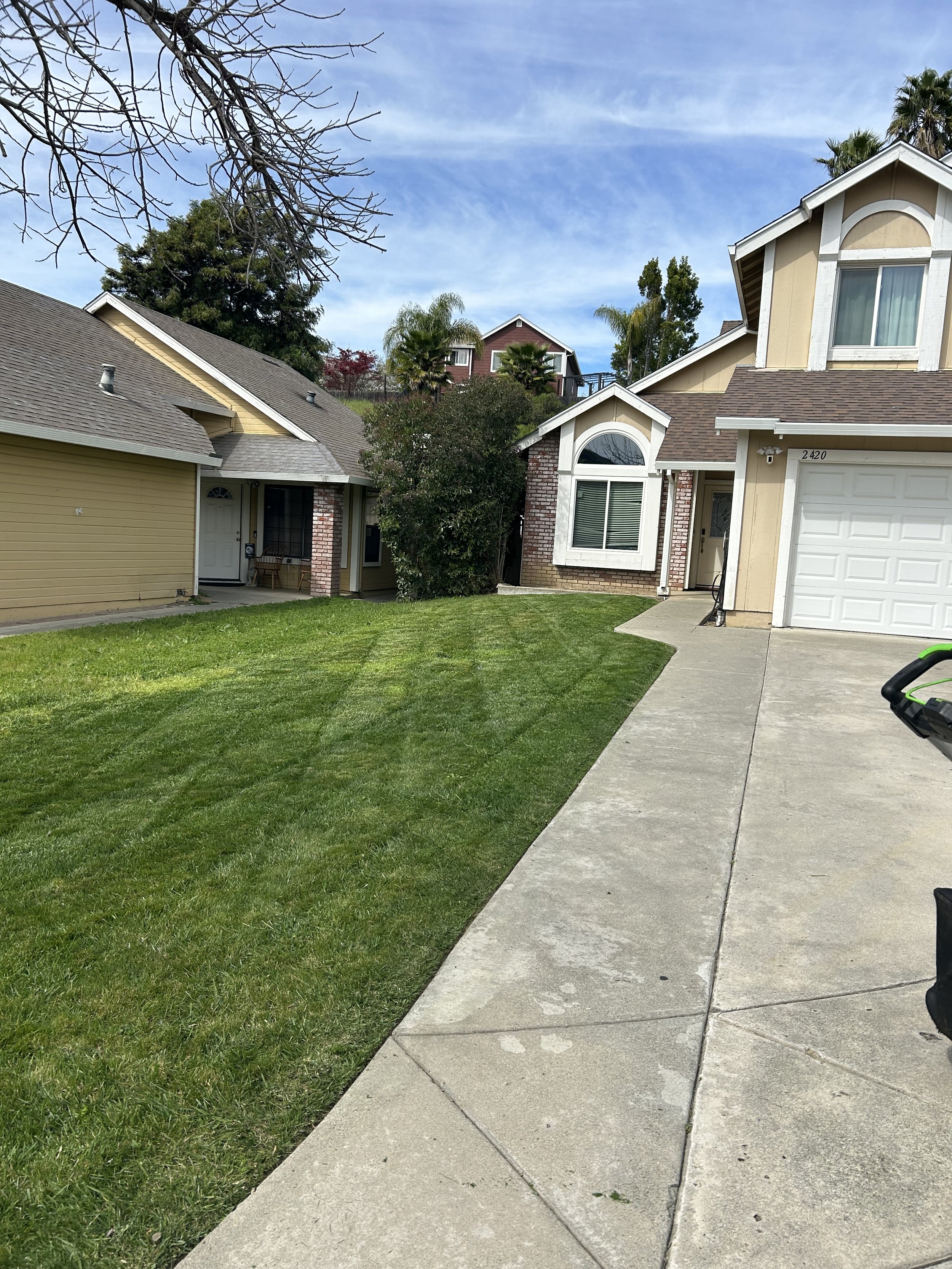 A suburban front yard with a green lawn, concrete driveway, and two attached houses with beige and yellow siding and white trim. Trees are visible in the background and the sky is partly cloudy.