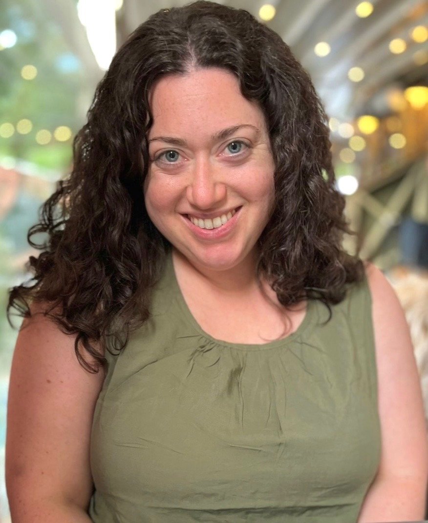 Emily Greenberger smiling at the camera, wearing a sleeveless olive green top, in a setting with blurred background lights.