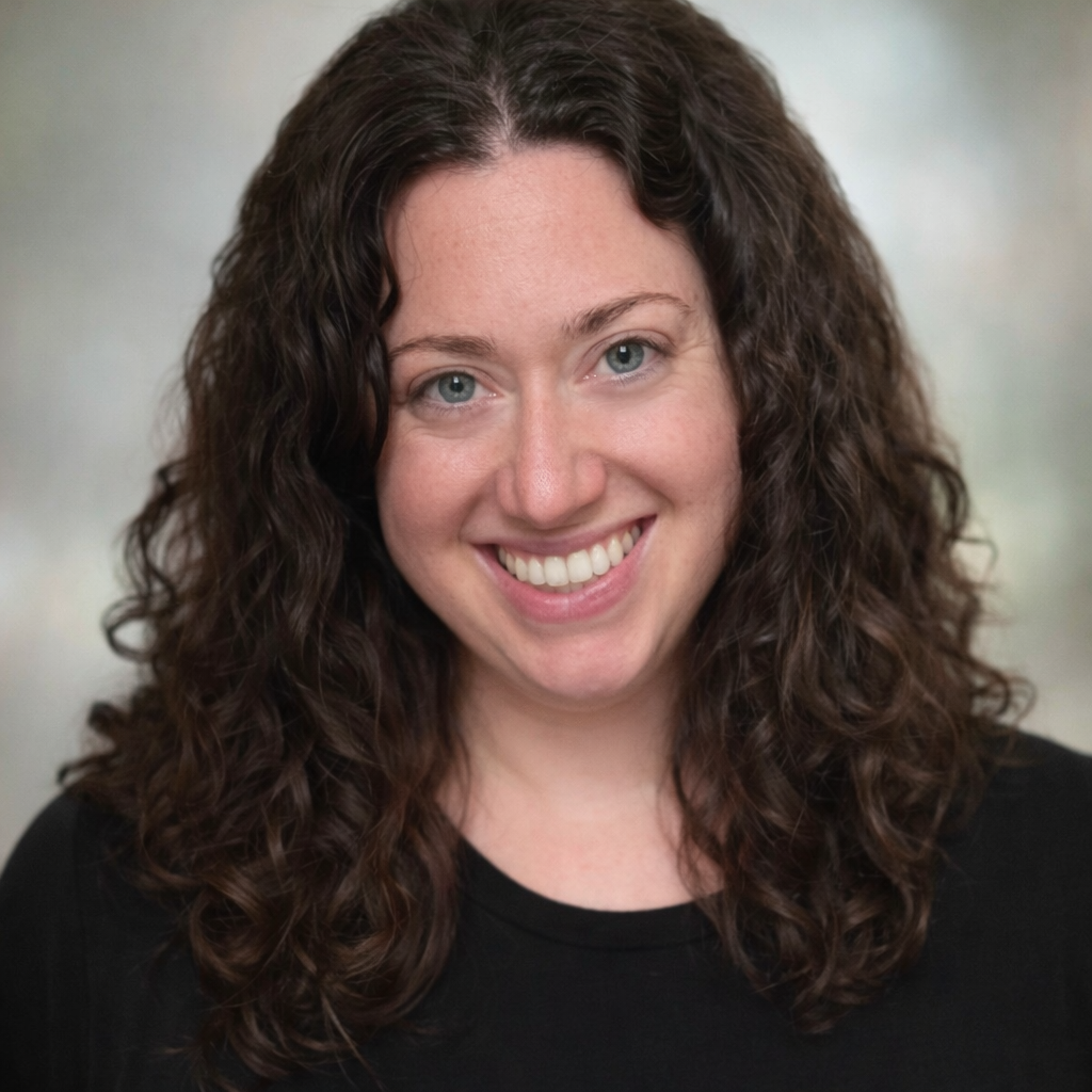 Emily Greenberger, smiling at the camera, wearing a black top, with a blurred background.