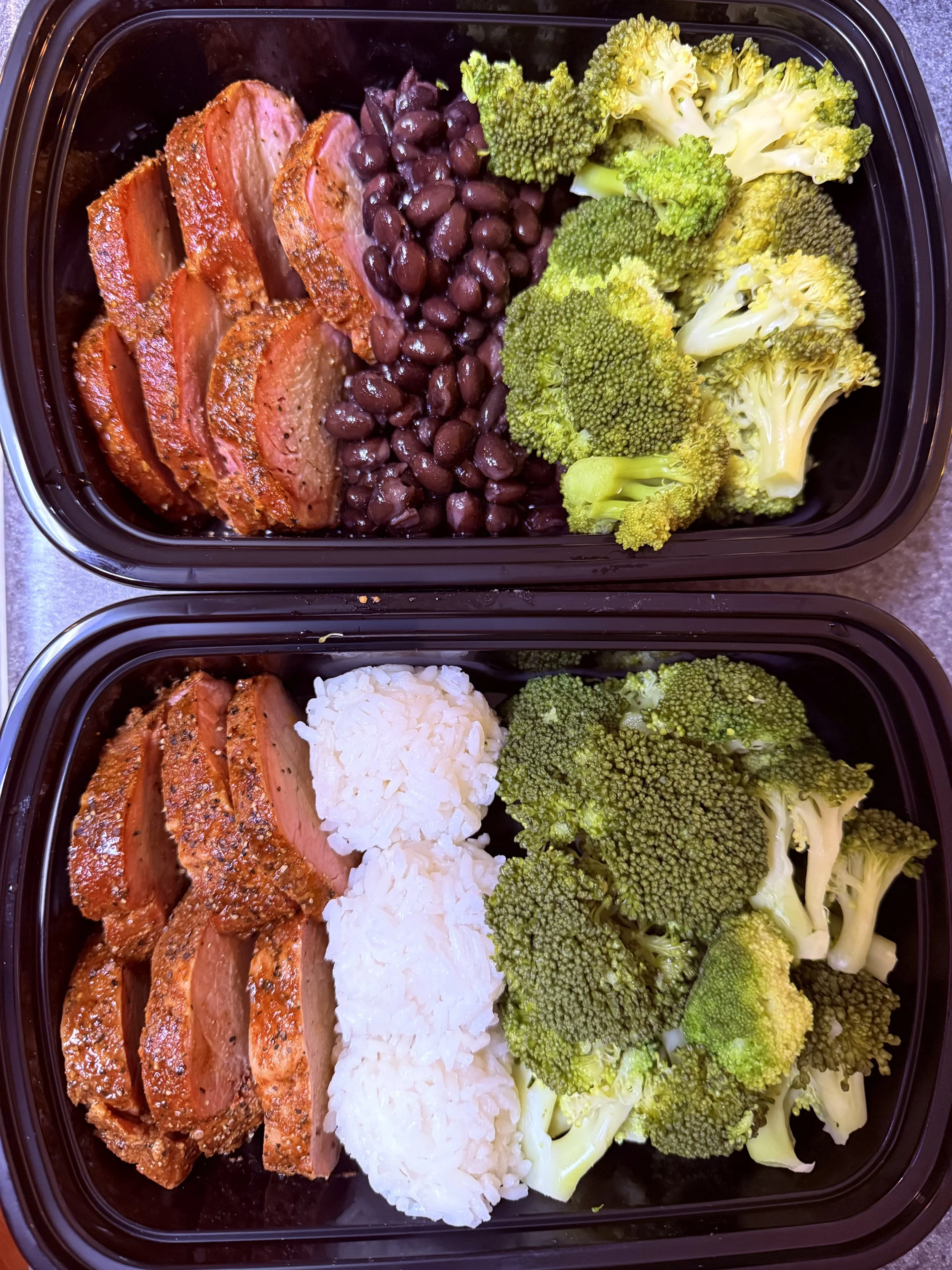 Two meal prep containers with sliced seasoned steak, black beans, steamed broccoli, and white rice.