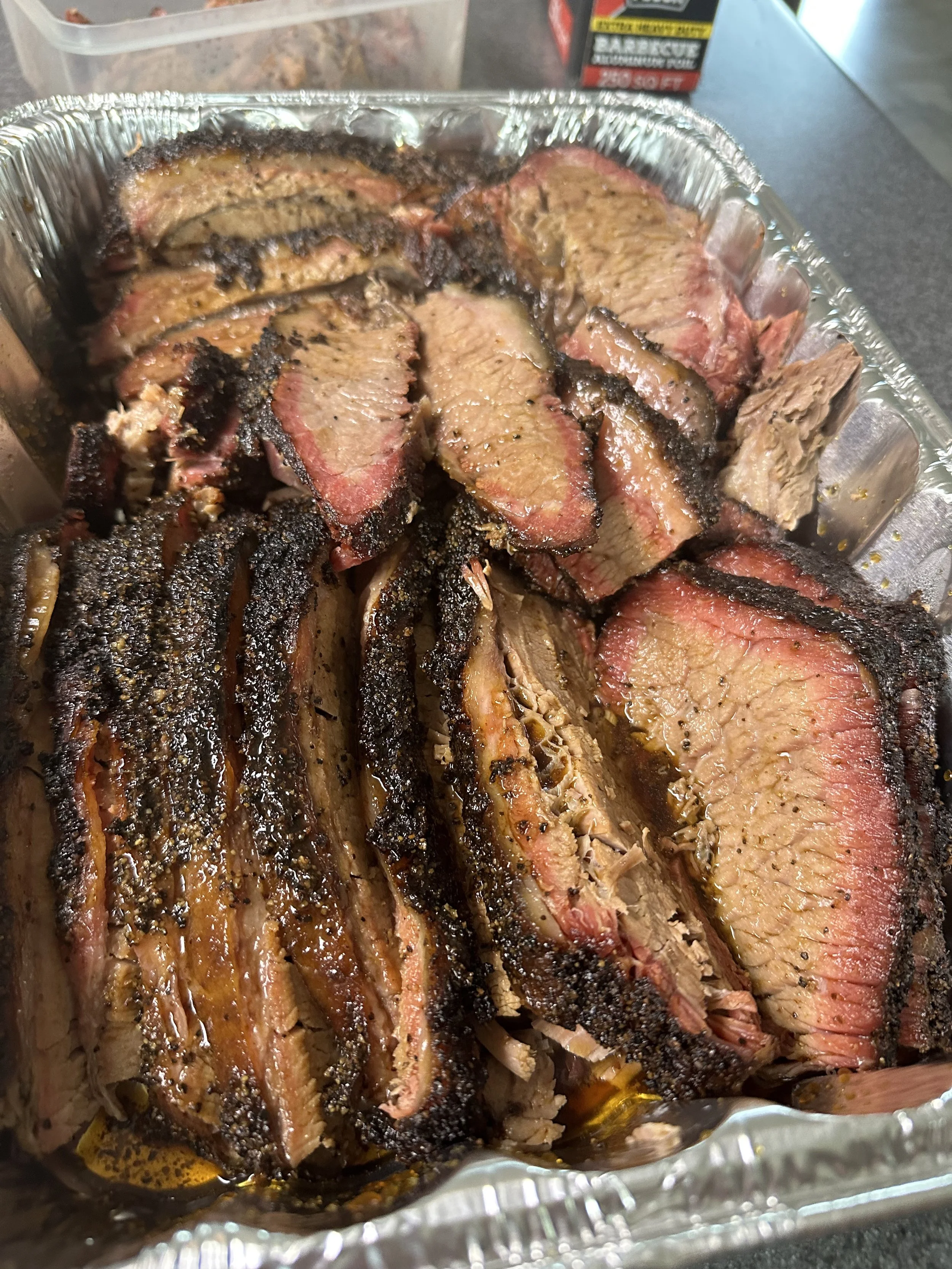Sliced smoked brisket in a foil tray with a dark bark crust and pink smoke ring.