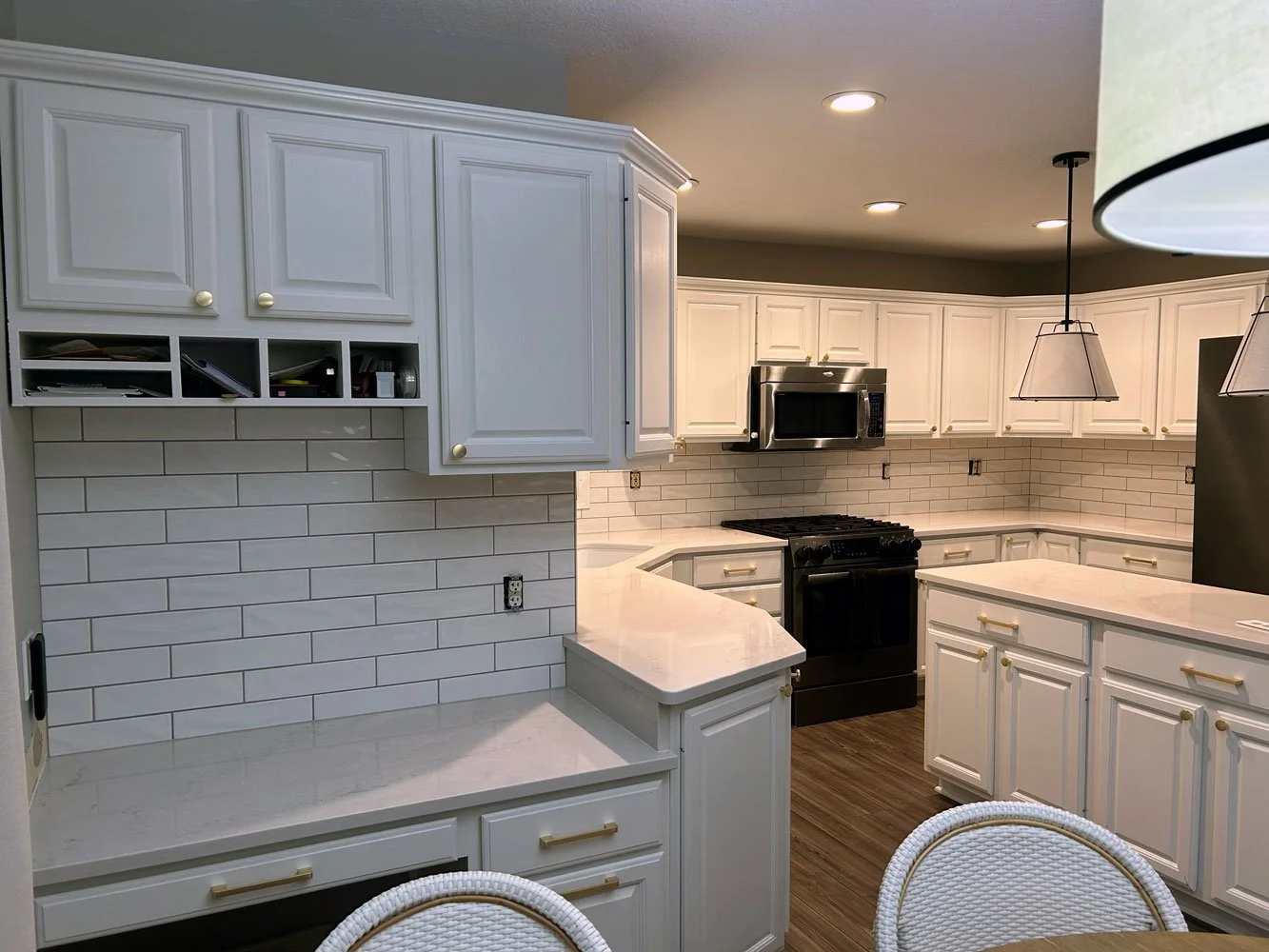 Kitchen with white cabinets, a white tile backsplash, black appliances including a microwave and a stove, and a white marble countertop. There are also pendant lights hanging from the ceiling and a wooden floor.