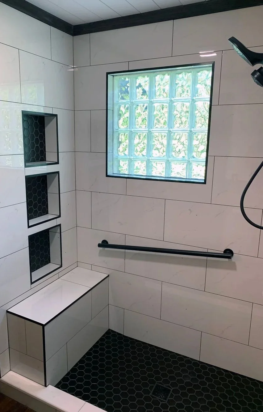 A modern shower interior with large white marble tiles, black hexagon floor tiles, a built-in black hexagon tray, a window made of glass blocks, black shelving, and a black grab bar on the wall.